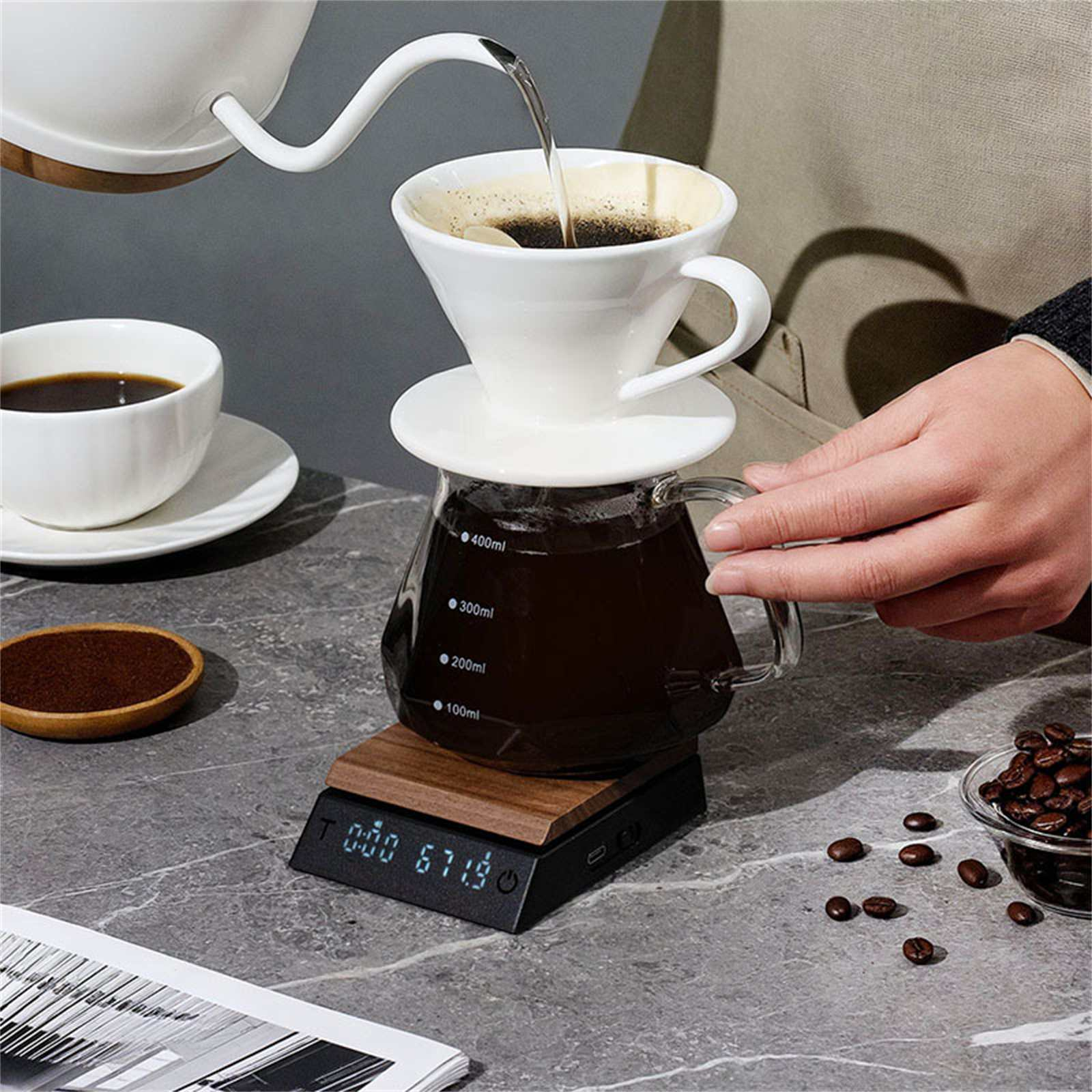 Kaffeezubereitung: Wasser gießt in einen Filter. Glaskaraffe auf einer Waage. Kaffeebohnen und eine Tasse daneben.