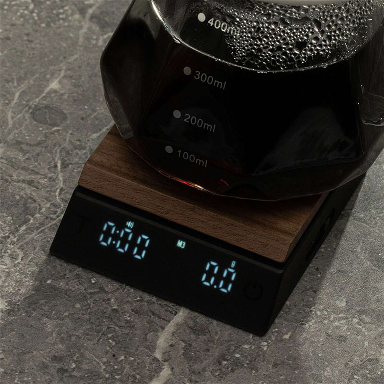 Kaffeezubereitung. Eine Glaskaraffe mit Flüssigkeit steht auf einer Waage, die Zeit und Gewicht anzeigt. Der Hintergrund ist eine Steinoberfläche.