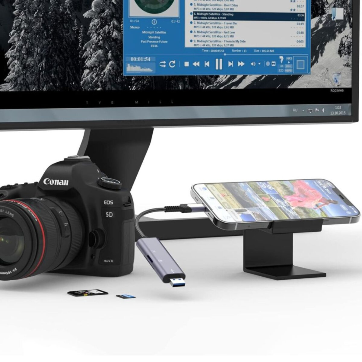 Una fotocamera DSLR, uno smartphone collegato a un hub e un monitor che mostra una playlist musicale.