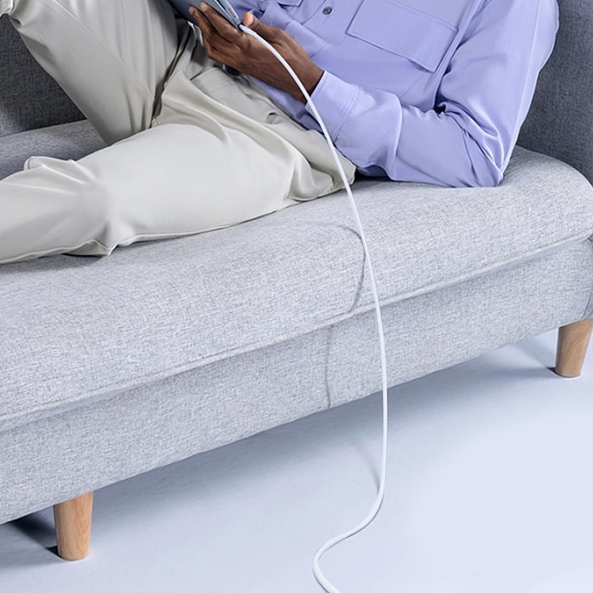 Person auf einem grauen Sofa, die ein Telefon benutzt, das mit einem weißen Ladekabel verbunden ist. Sie trägt ein lavendelfarbenes Hemd und beige Hosen.