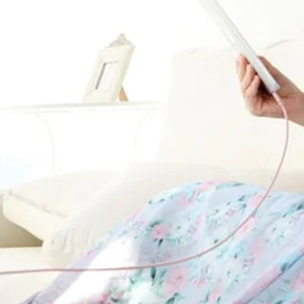 Eine Person hält ein Tablet mit pinkfarbenem Kabel. Eine Blumendecke, ein Kissen und ein Bilderrahmen sind sichtbar. Die Szene spielt sich drinnen ab.
