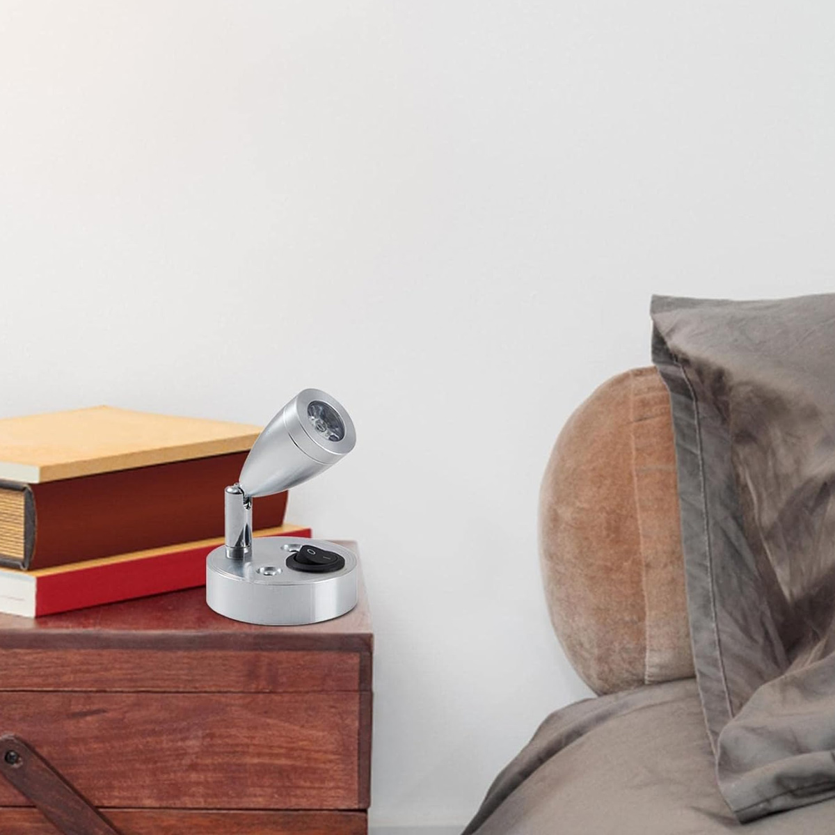 Eine silberne Nachttischlampe steht auf einem hölzernen Nachttisch mit Büchern. Rechts befindet sich ein Bett mit braunem Kopfteil und grauer Bettwäsche.