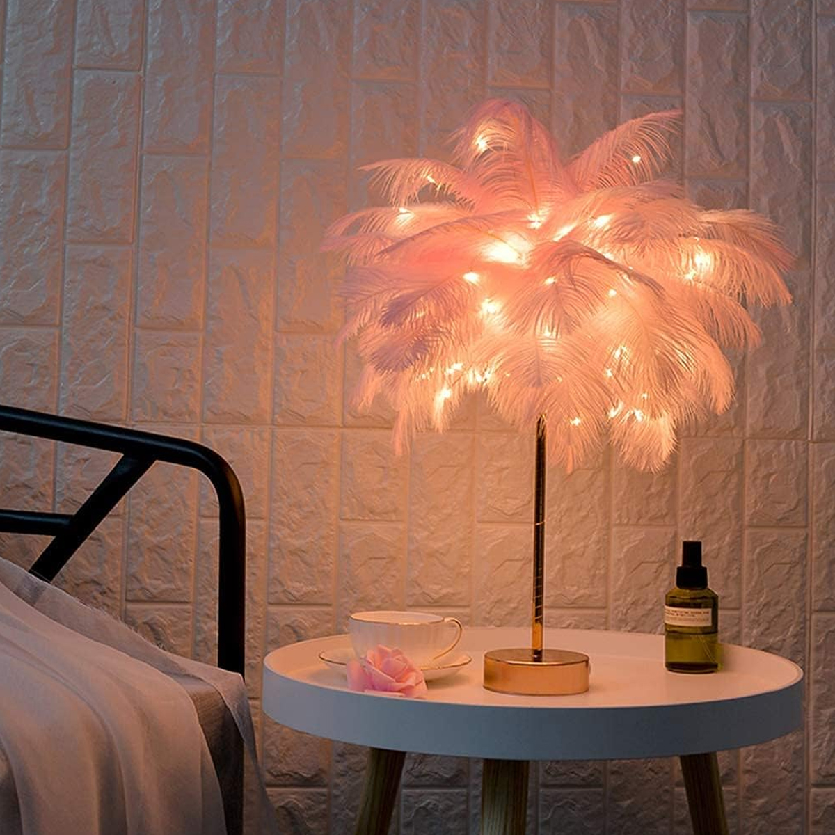 Eine dekorative Lampe mit rosa Federn und Lichtern auf einem weißen Tisch. Eine Tasse, eine Rose und eine Flasche stehen ebenfalls auf dem Tisch.