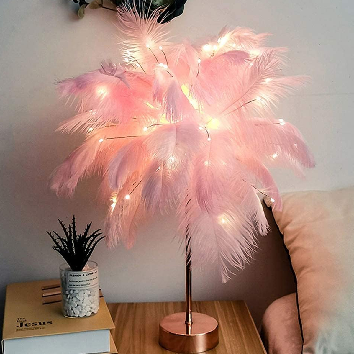 Eine rosa Federlampe mit Lichterketten steht auf einem Nachttisch. Eine kleine Pflanze und ein Buch sind ebenfalls auf dem Tisch.