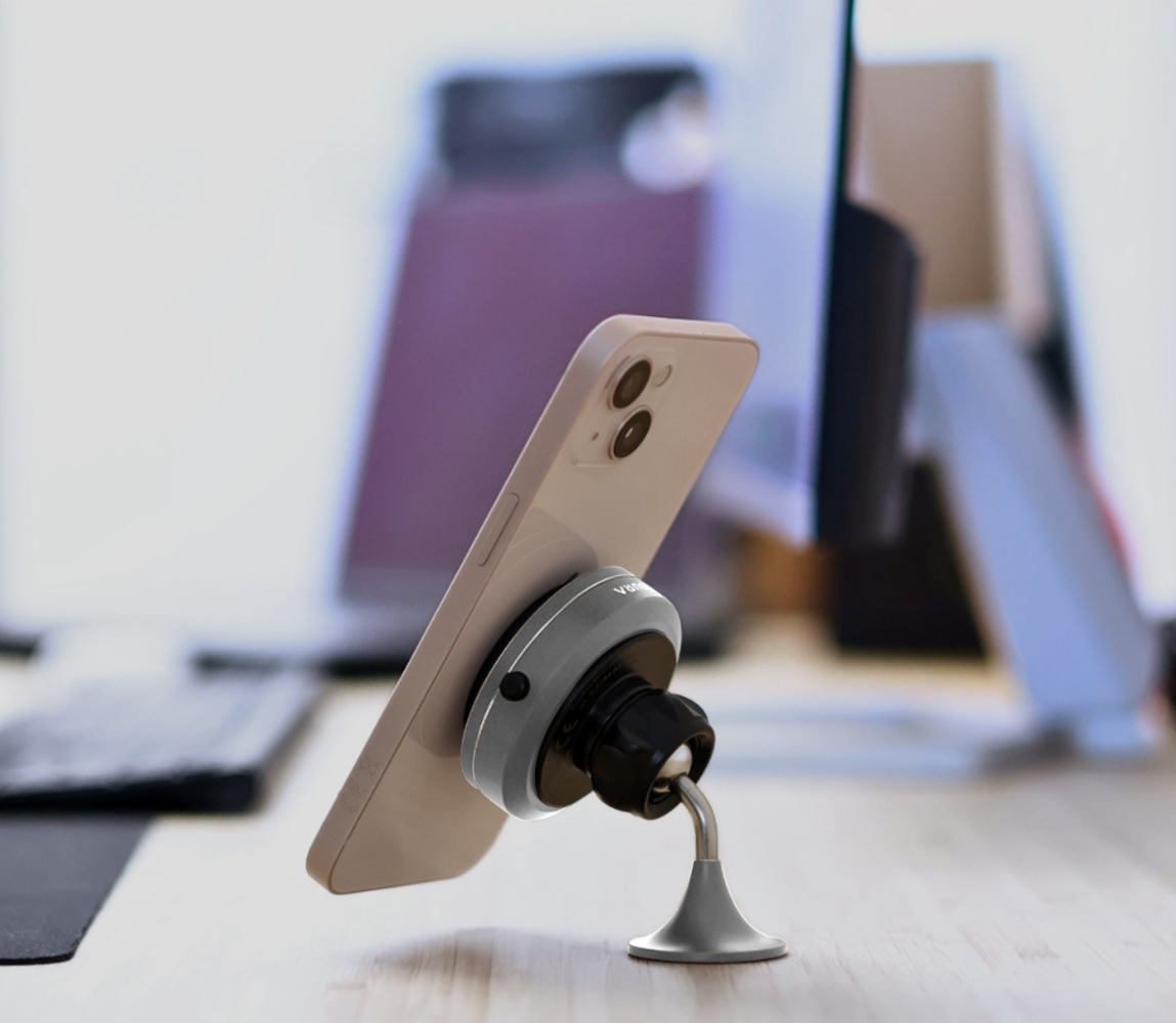 Uno smartphone beige è montato su un supporto grigio. Un computer e una tastiera sono sfocati sullo sfondo su una scrivania di legno.
