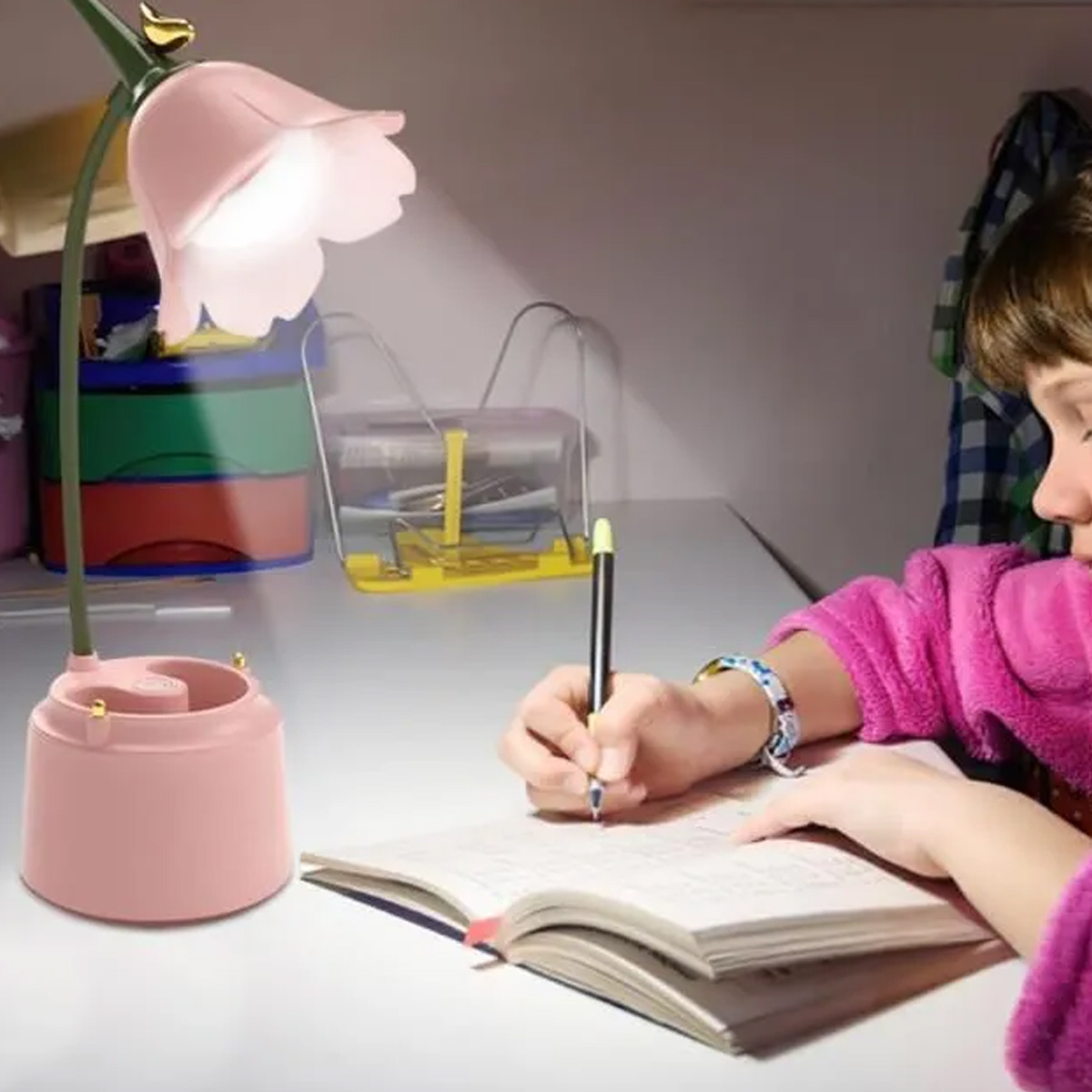 Ein Mädchen schreibt in ein Notizbuch, beleuchtet von einer rosa blütenförmigen Lampe. Im Hintergrund befindet sich ein Schreibtisch mit Schreibwaren.
