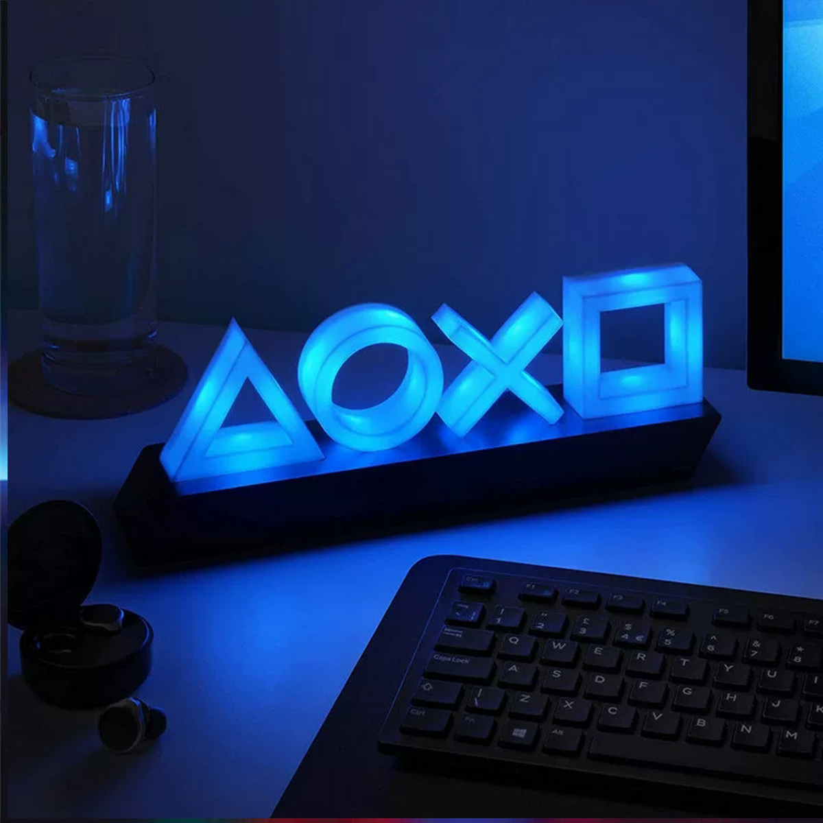 Ein blau leuchtendes PlayStation-Symbol-Licht steht auf einem Schreibtisch mit einer Tastatur, Ohrhörern und einem Glas Wasser.