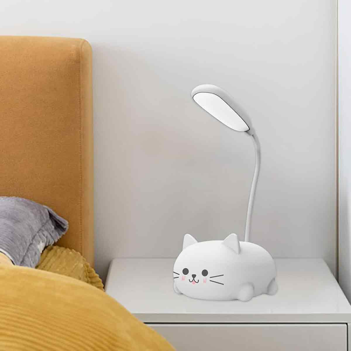 Eine weiße, katzenförmige Schreibtischlampe steht auf einem weißen Tisch. Im Hintergrund ist ein Bett. Die Lampe ist an.