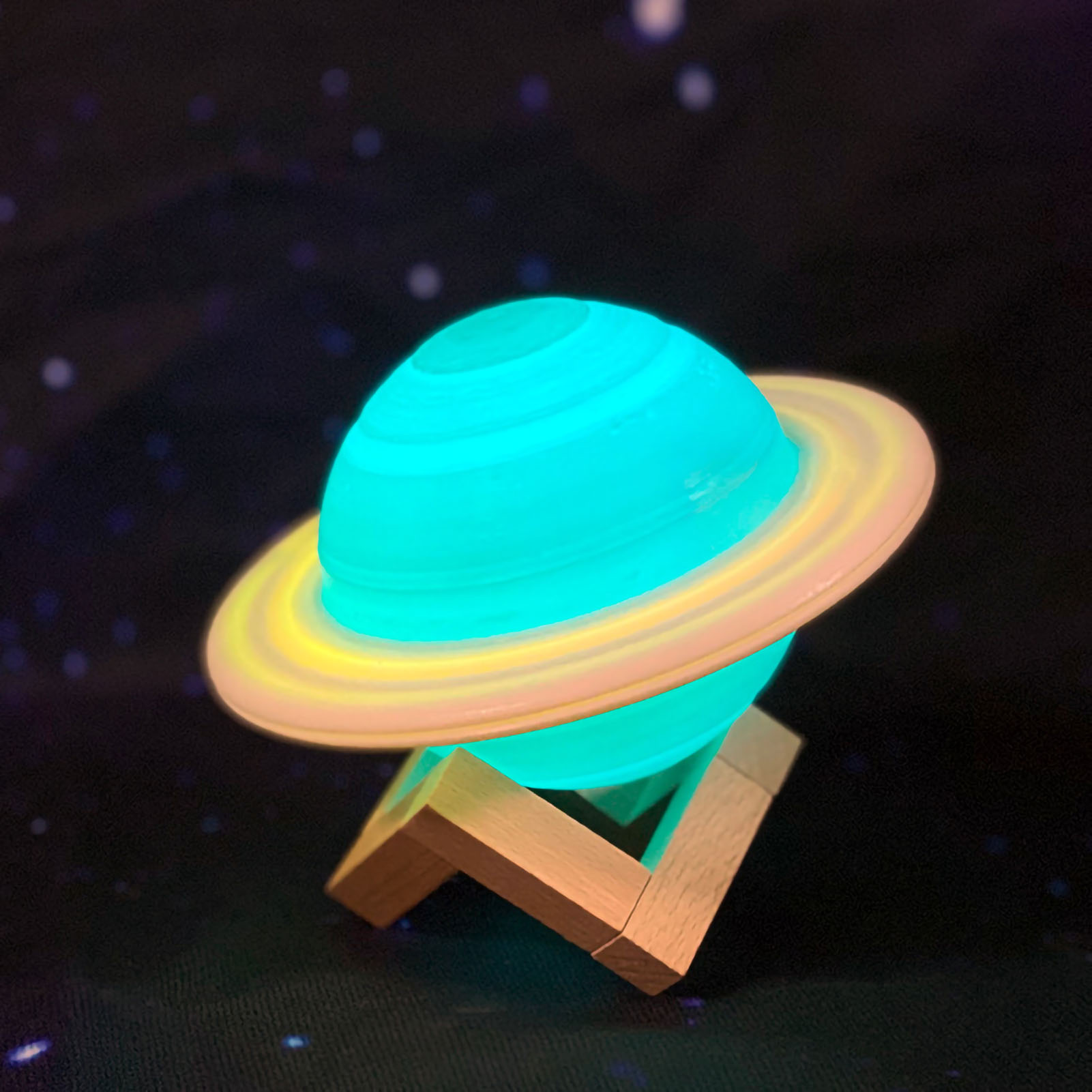 Eine leuchtende Planetenlampe mit Ringen, türkisfarbenem Körper und gelb-weißen Ringen, auf einem Holzständer, vor Sternenhimmel.