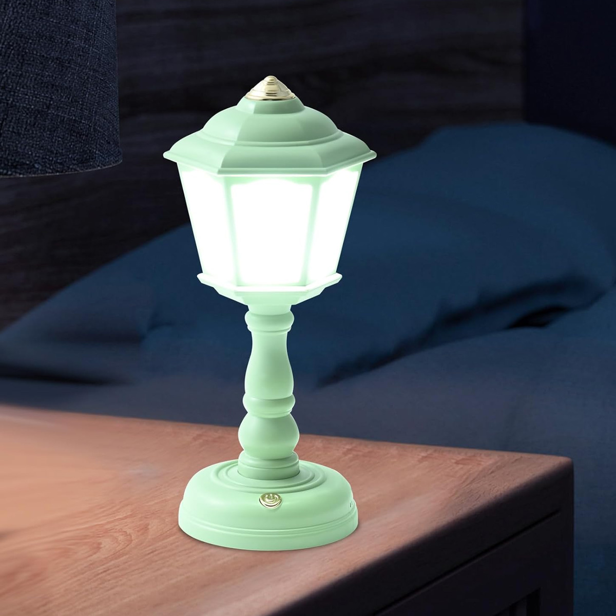 Eine mintgrüne laternenförmige Lampe mit goldenem Dach auf einem Holztisch. Im Hintergrund ein Bett.