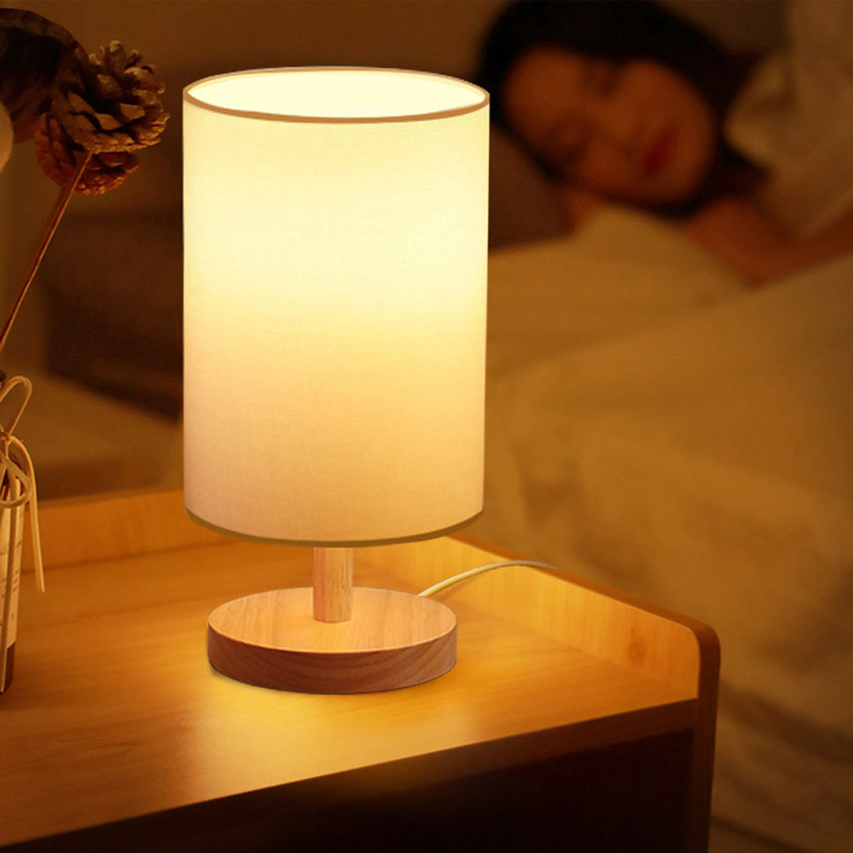 Eine beleuchtete Nachttischlampe auf einem Holztisch. Im Hintergrund schläft eine Person. Eine Kiefernzapfen und Blumen sind an der Seite.