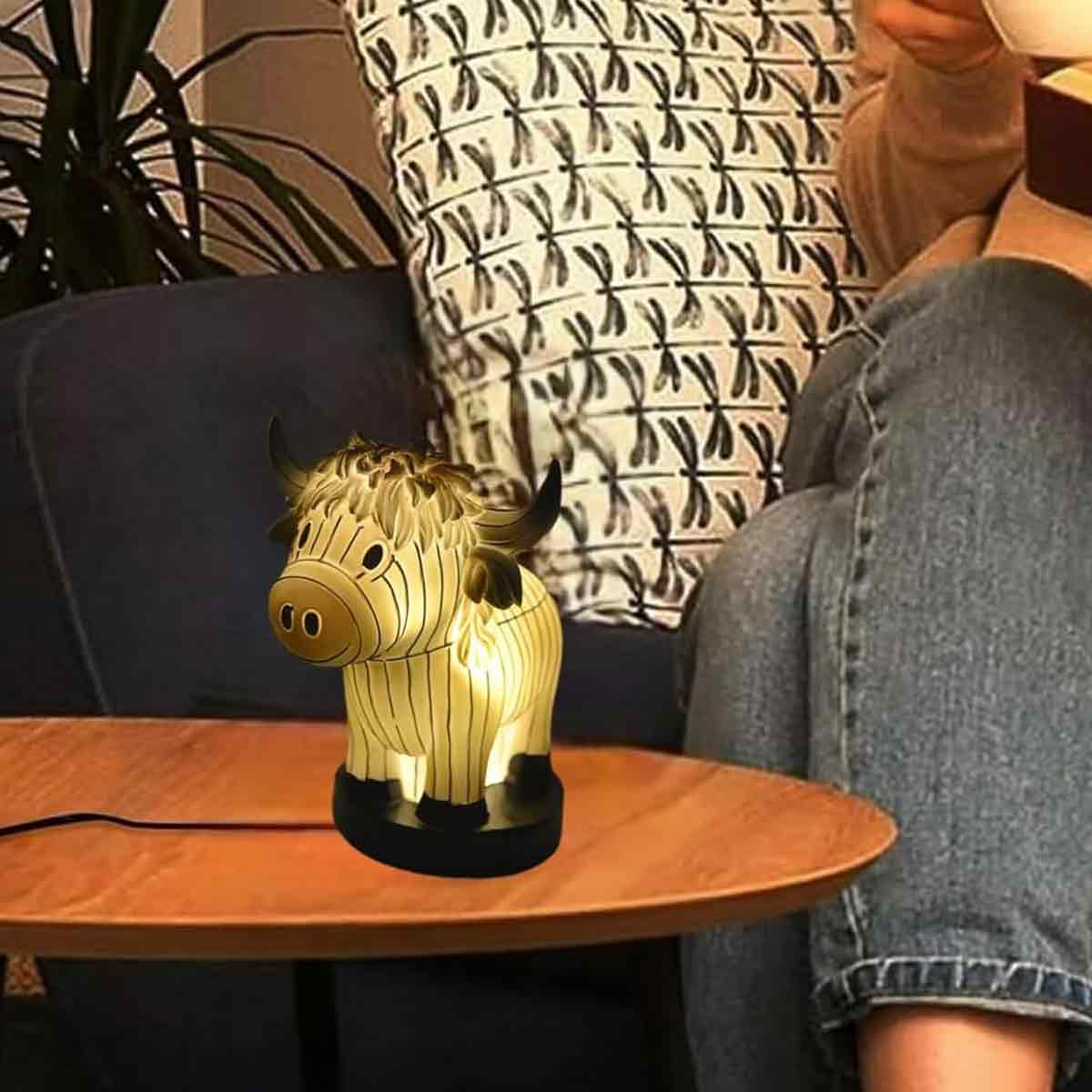 Eine beleuchtete Kuhlampe auf einem Tisch, mit einer Person im Hintergrund. Die Lampe steht auf einem Holztisch. Ein gemustertes Kissen ist dahinter.