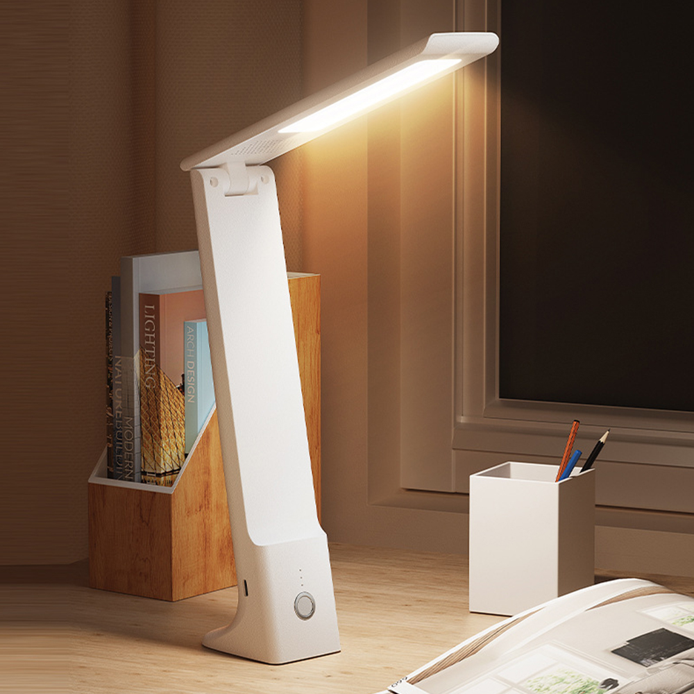 Eine weiße Schreibtischlampe leuchtet und beleuchtet den Schreibtisch. Bücher, Bleistifte und eine Zeitschrift sind sichtbar. Im Hintergrund ist ein Fenster.