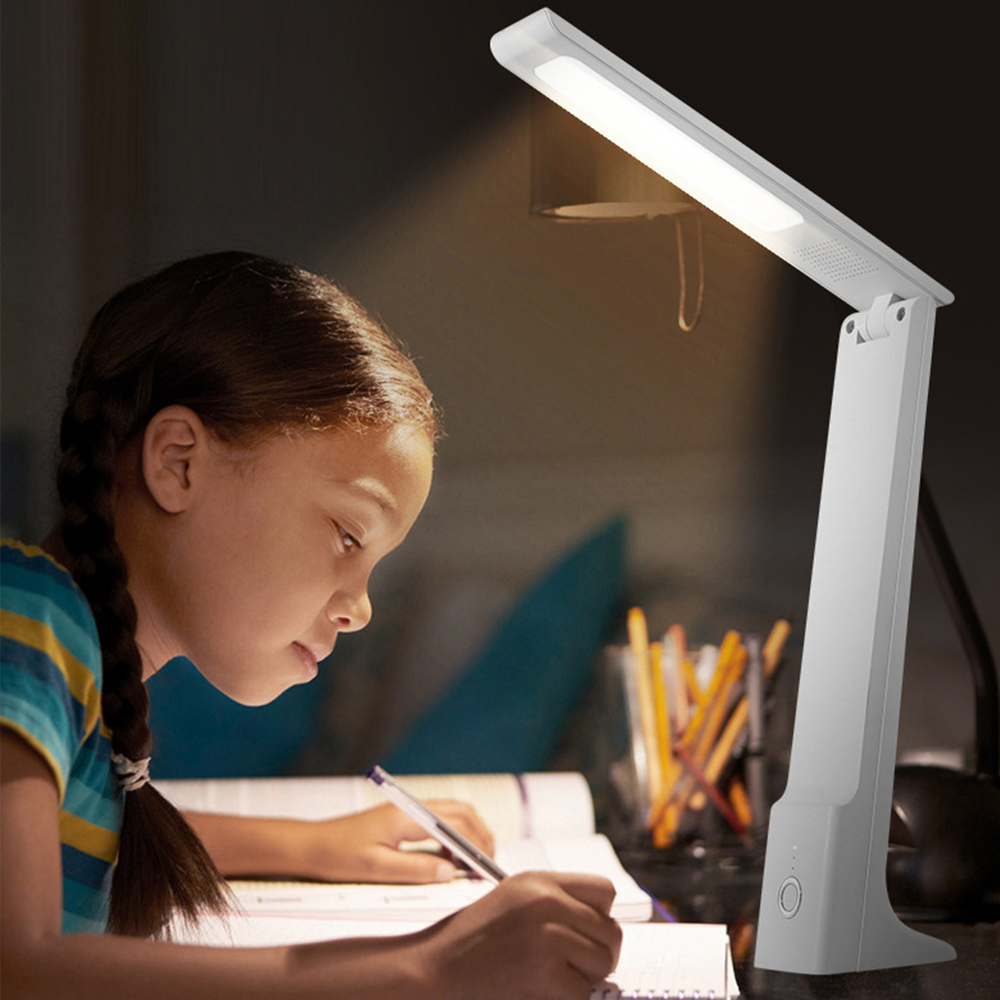 Ein Mädchen schreibt an einem Schreibtisch, der von einer weißen Schreibtischlampe beleuchtet wird. Sie trägt ein gestreiftes Hemd. Auf dem Schreibtisch liegen ein Notizbuch und Stifte.
