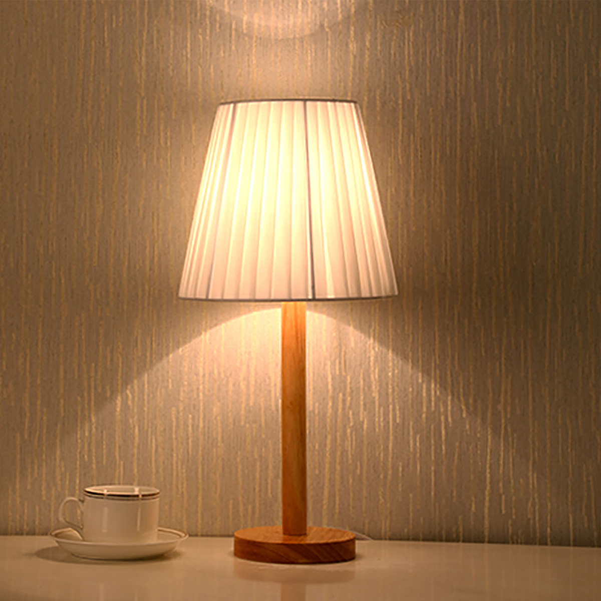 Eine beleuchtete Lampe mit weißem Lampenschirm steht auf einem Tisch neben einer Teetasse. Der Hintergrund ist eine strukturierte Wand.