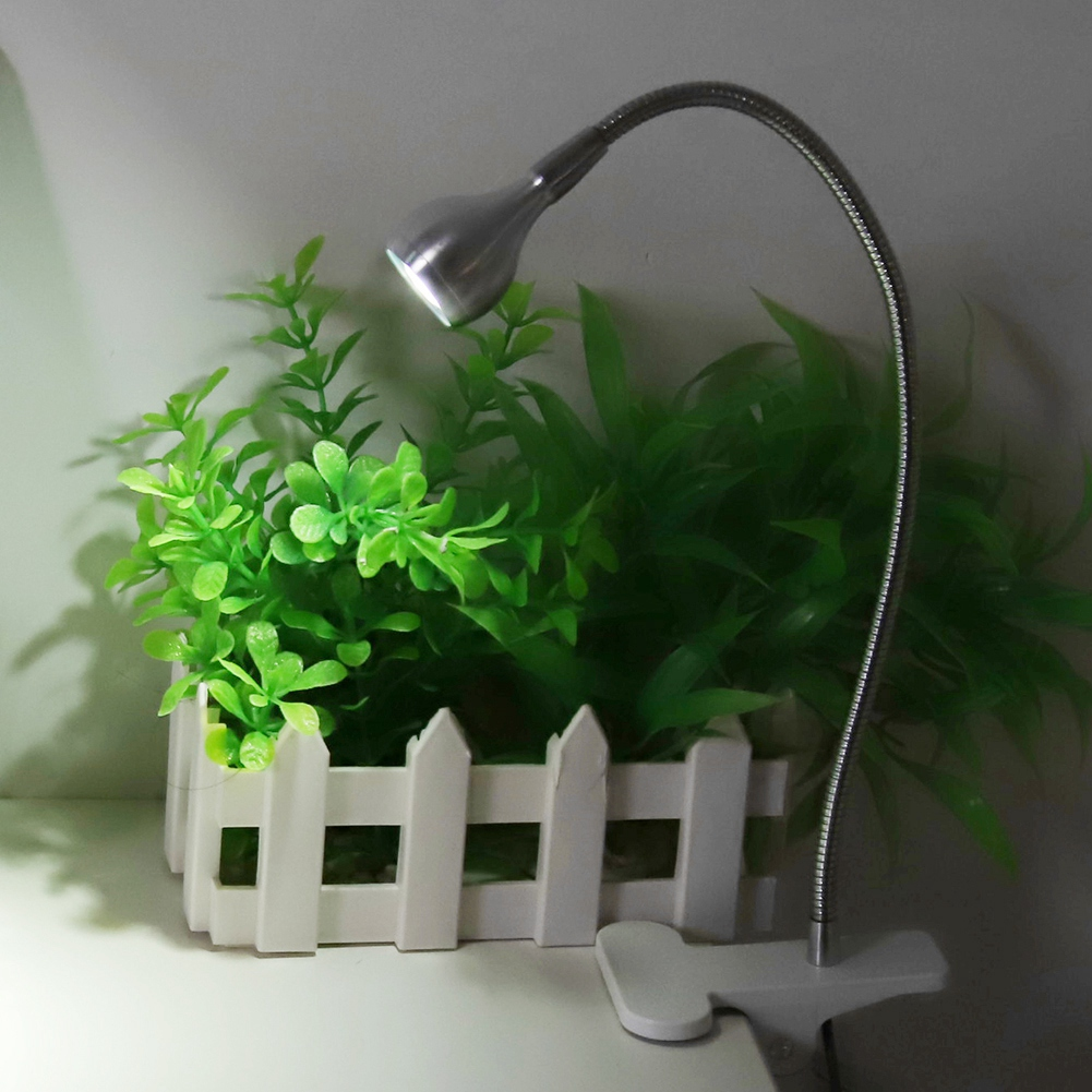 Eine Schreibtischlampe mit silbernem Kopf und flexiblem Arm ist an einem Tisch befestigt. Grüne Pflanzen hinter einem weißen Zaun.