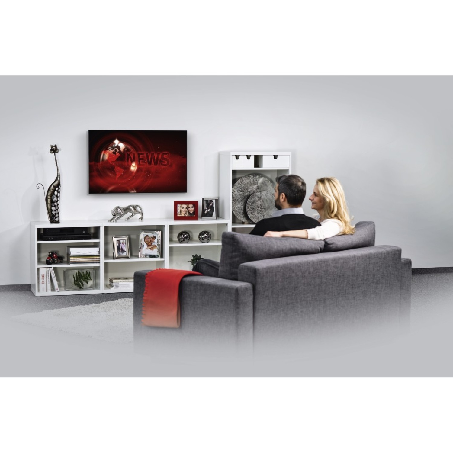 Ein Paar sitzt auf einem grauen Sofa und schaut Nachrichten im Fernsehen. Ein weißer Schrank und Dekorationsgegenstände sind links.