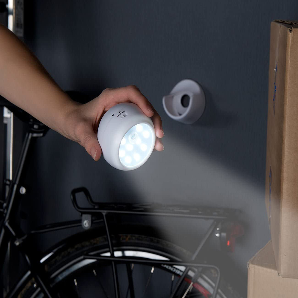 Eine Hand hält eine leuchtende, weiße LED-Leuchte. Im Hintergrund sind ein Fahrrad, ein Halter und Kartons zu sehen.