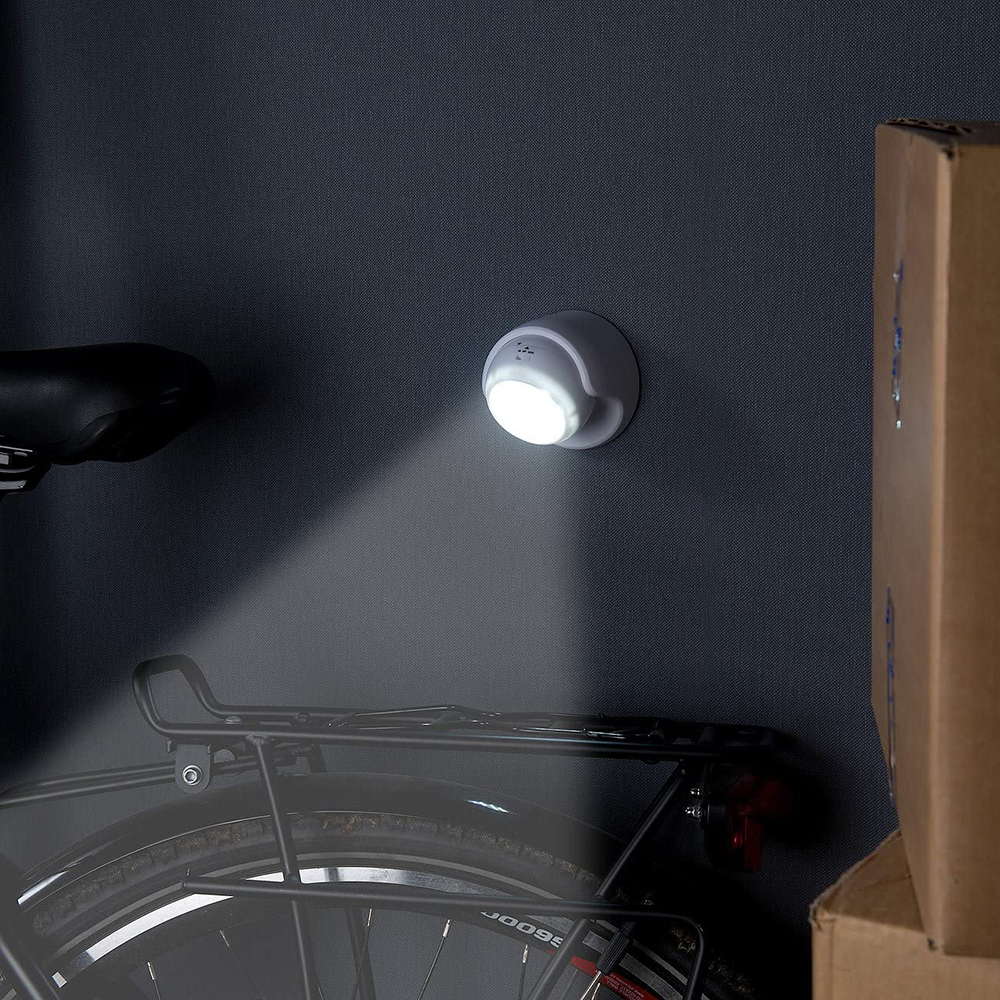 Eine weiße LED-Leuchte scheint auf eine Wand. Darunter ist ein Fahrrad, rechts ein Karton.