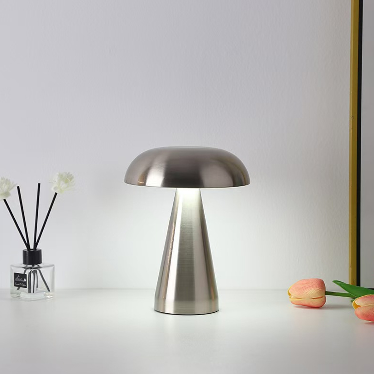 Eine metallische, pilzförmige Lampe steht auf einer weißen Oberfläche. Blumen und ein Diffusor links, Tulpen rechts.