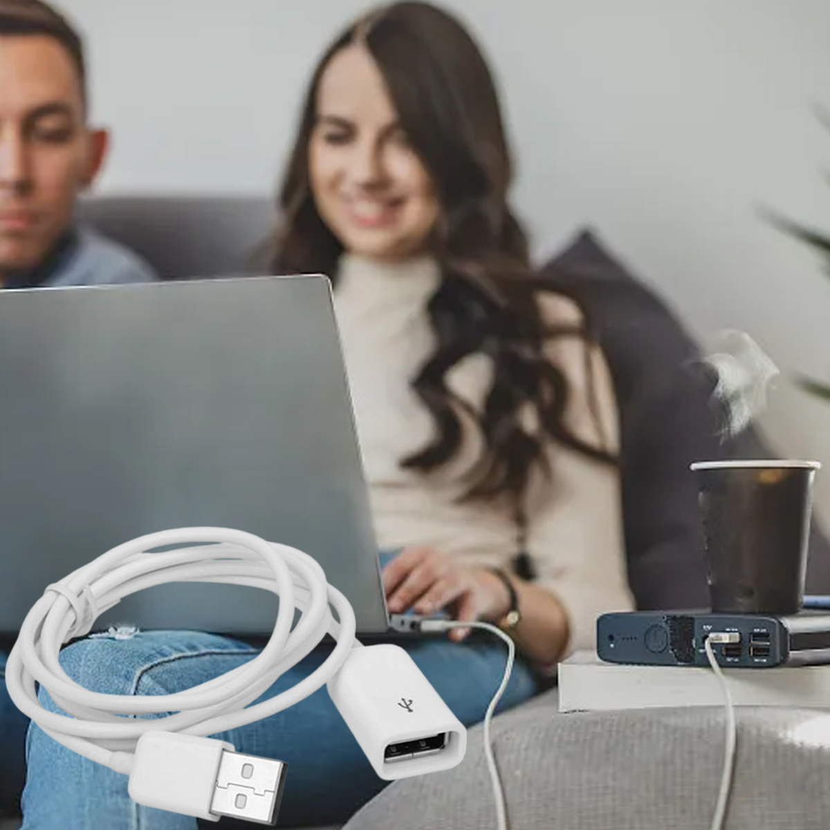 Ein weißes USB-Kabel und ein Adapter sind im Vordergrund. Zwei Personen im Hintergrund benutzen einen Laptop. Rechts eine Powerbank.