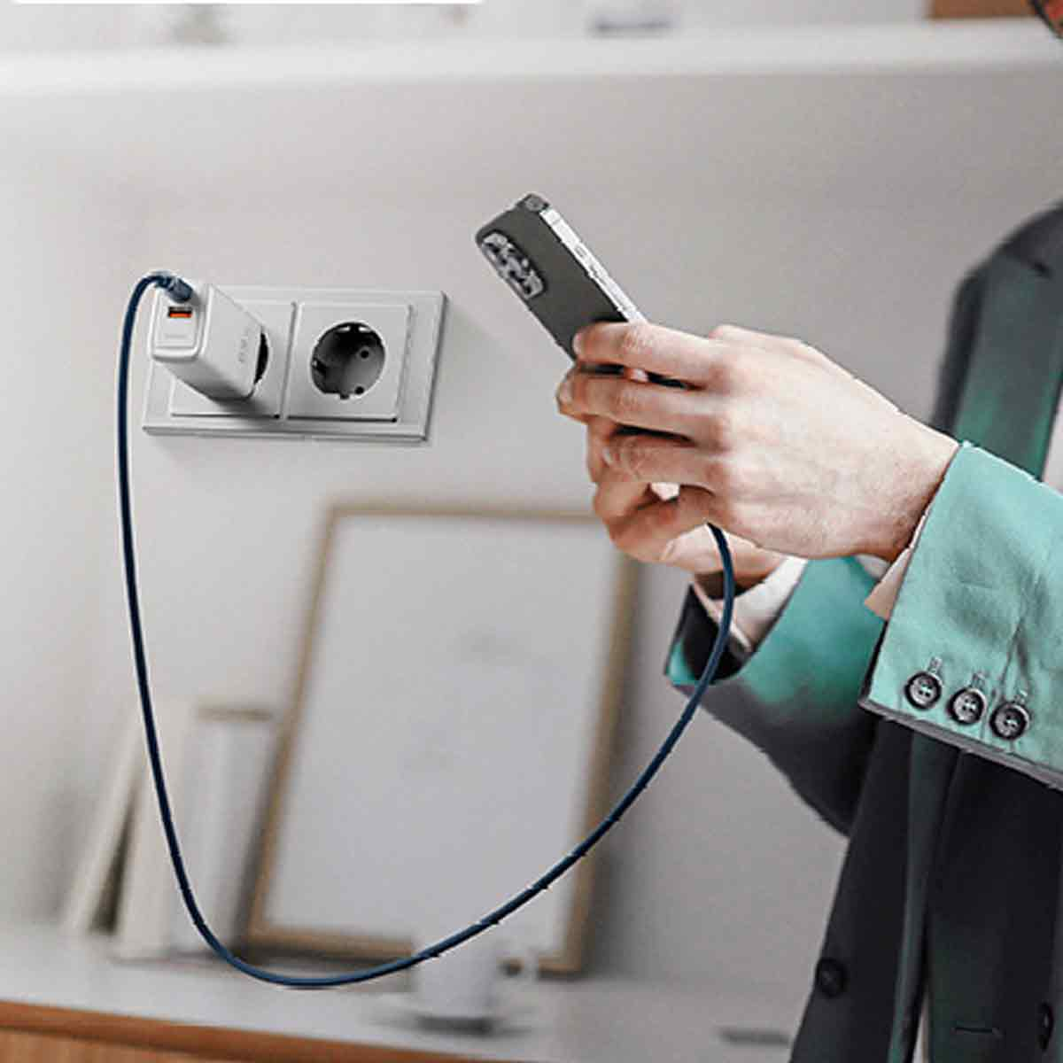 Eine Person hält ein Telefon, das mit einem Ladegerät verbunden ist, das an eine Steckdose angeschlossen ist. Die Person trägt einen grünen Blazer.