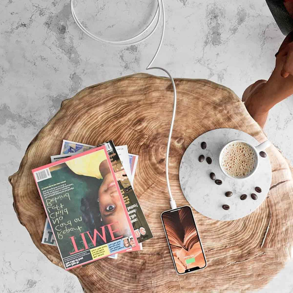 Ein Telefon, das auf einem Holztisch geladen wird, mit einer Kaffeetasse, Zeitschriften und Kaffeebohnen. Der Hintergrund ist eine Marmoroberfläche.