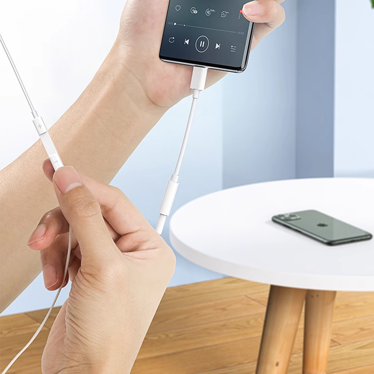 Eine Hand hält ein Telefon mit weißem Adapter. Eine andere Hand hält das Kopfhörerkabel. Ein Telefon liegt auf einem Tisch.
