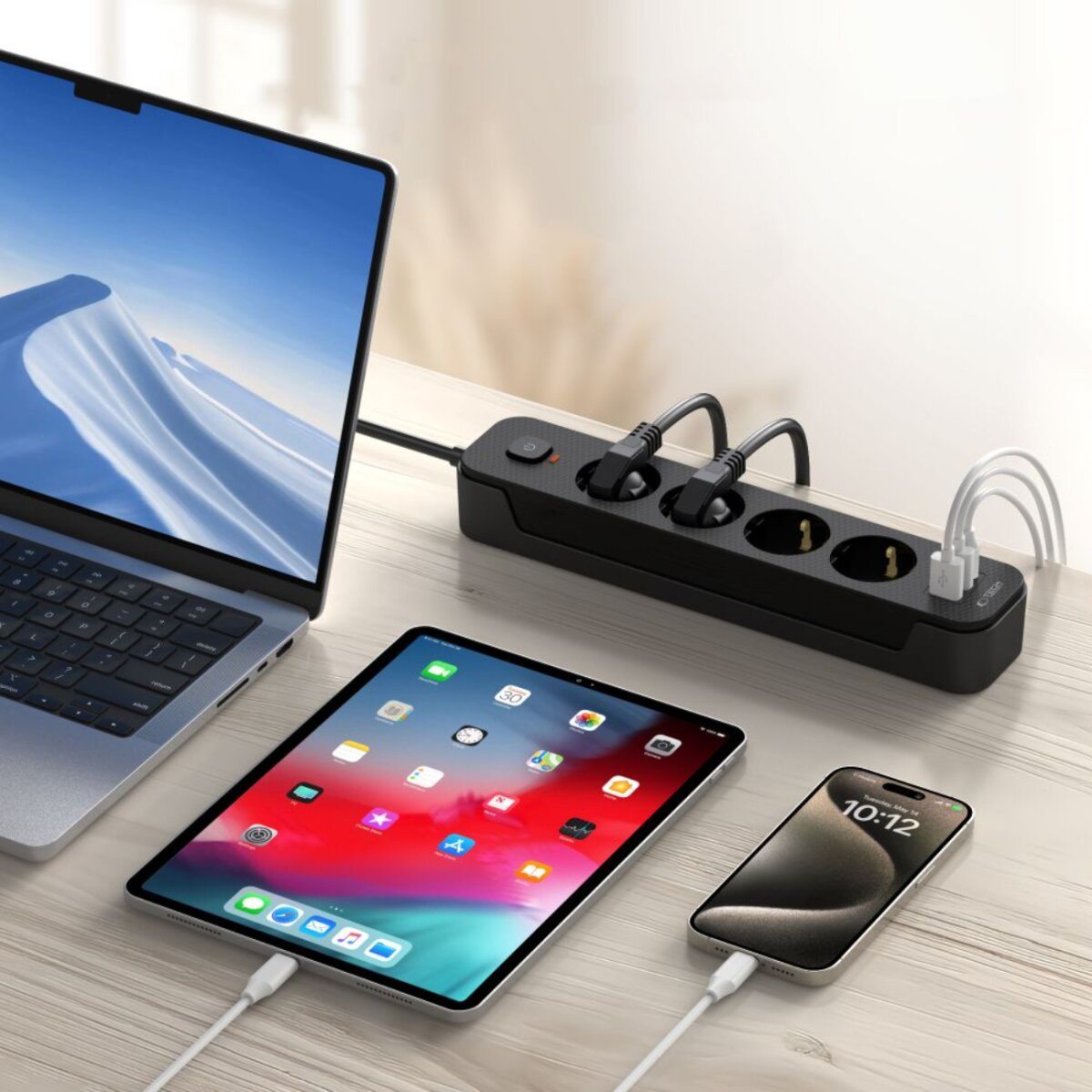 Ein Laptop, ein Tablet und ein Telefon laden auf einem Holztisch. Eine schwarze Steckdosenleiste mit mehreren Steckdosen ist zu sehen.