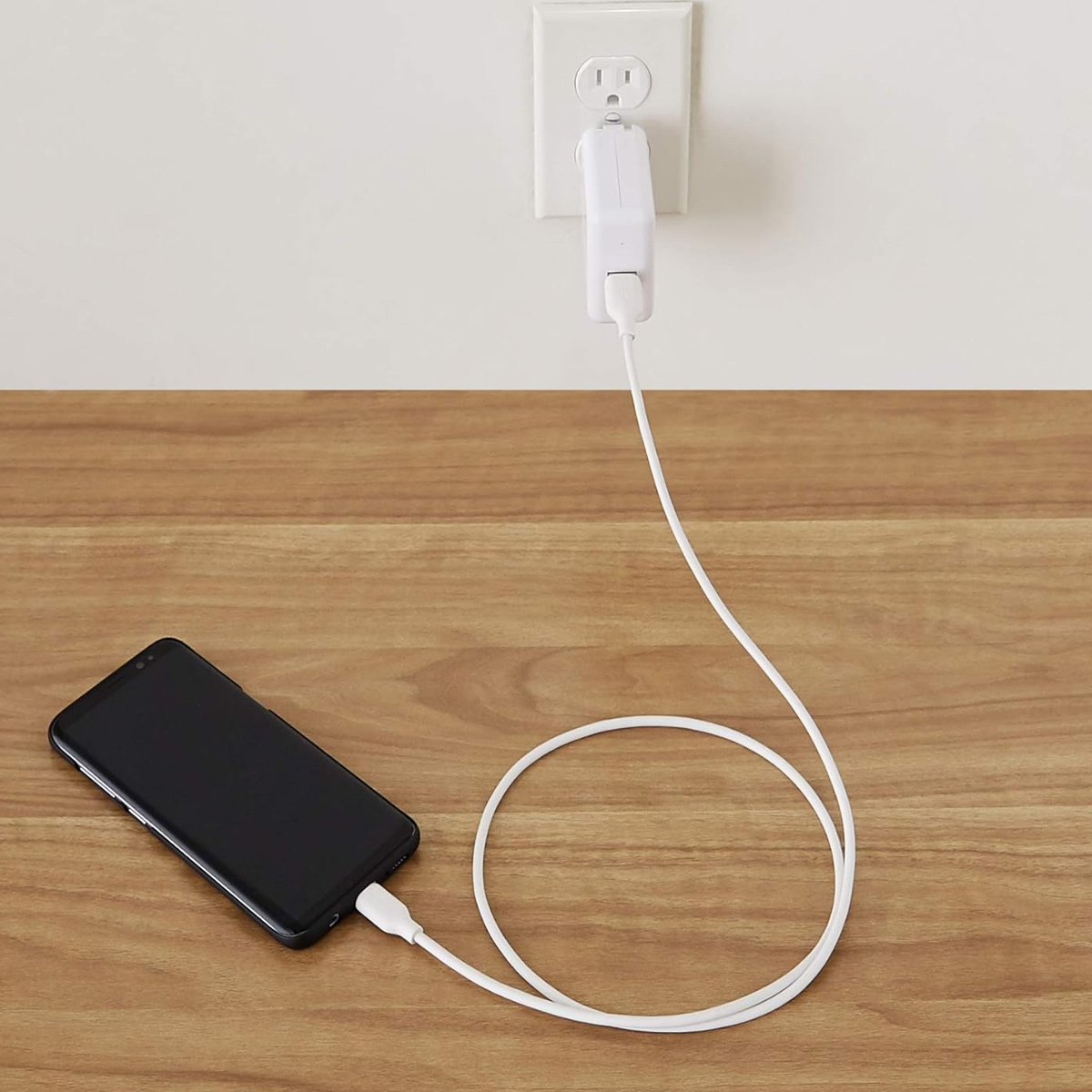 Ein schwarzes Smartphone wird auf einer Holzoberfläche aufgeladen. Ein weißes Kabel ist an einer Steckdose angeschlossen.