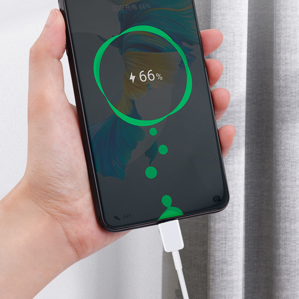 Hand hält ein ladendes Telefon. Der Bildschirm zeigt 66 % Ladung mit einem grünen Kreis. Weißes Kabel angeschlossen. Grauer Vorhang im Hintergrund.