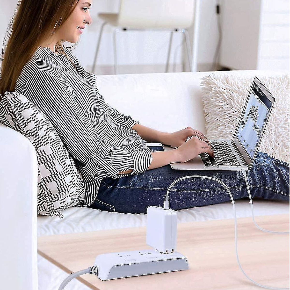 Eine Frau sitzt auf einem Sofa und benutzt einen Laptop. Eine Steckdosenleiste und ein Ladegerät stehen auf einem Tisch. Sie trägt ein gestreiftes Hemd und Jeans.