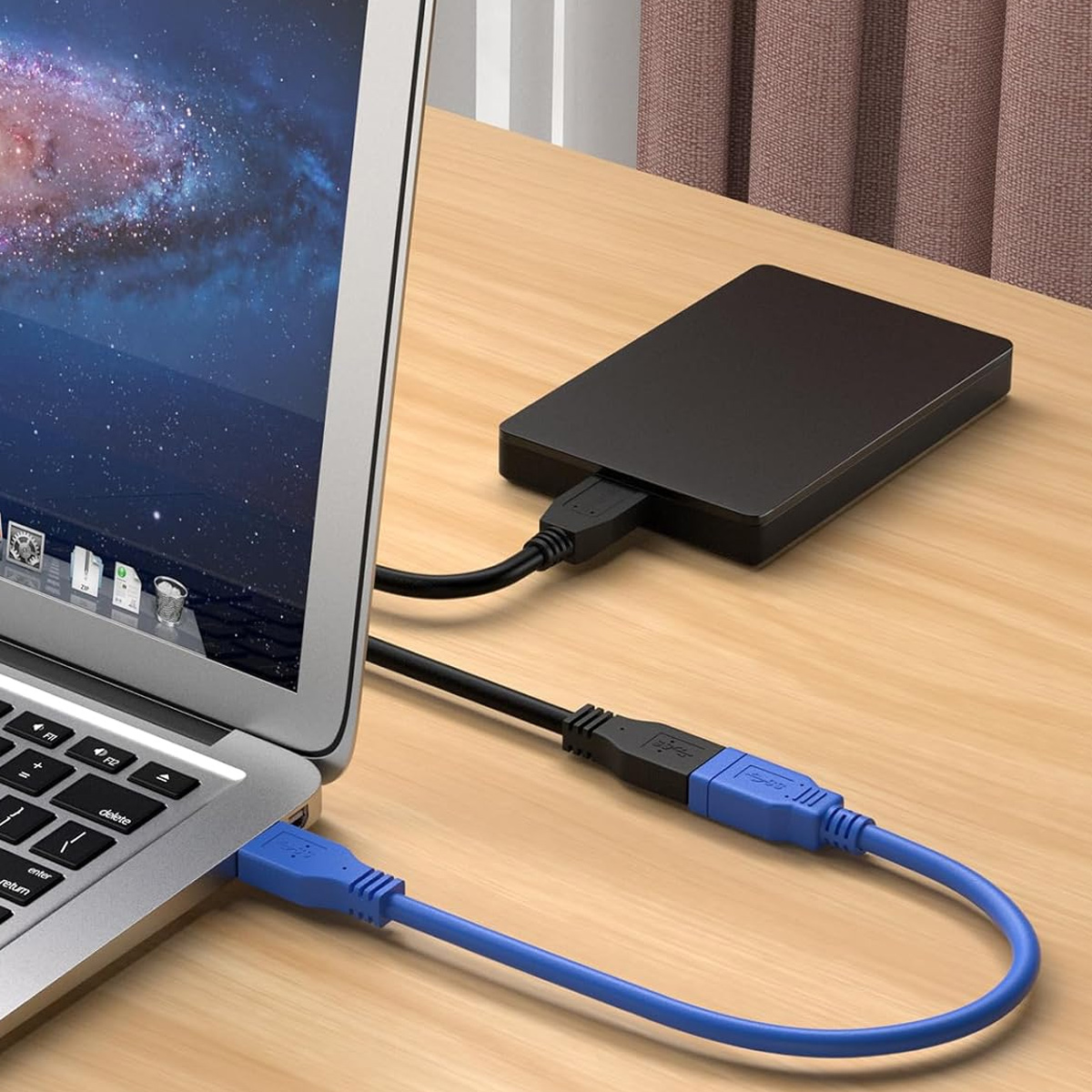 Ein Laptop, der über blaue und schwarze USB-Kabel mit einer schwarzen externen Festplatte verbunden ist, auf einem Holztisch.