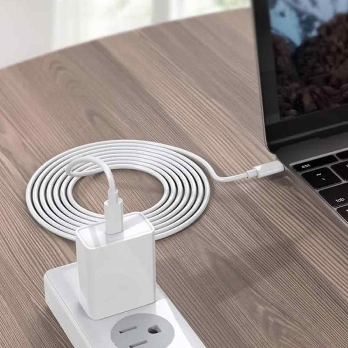 Ein weißes Laptop-Ladegerät ist an einen Laptop und eine Steckdose angeschlossen. Das Kabel ist auf einer Holzoberfläche aufgerollt.