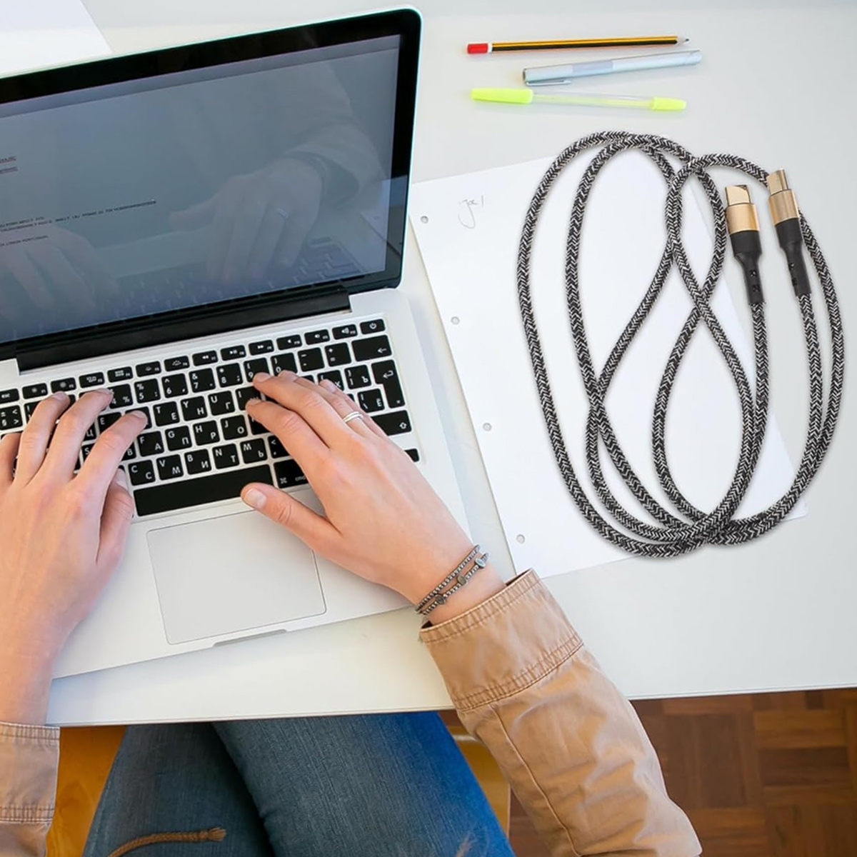 Eine Person tippt auf einem Laptop. Kabel und Stifte liegen auf dem Tisch. Die Person trägt eine Jacke und ein Armband.