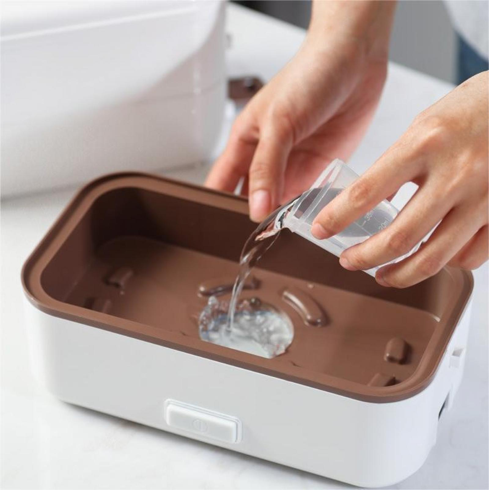 Hände gießen Wasser aus einem Messbecher in einen braun-weißen Essensbehälter. Ein weißer Behälter ist im Hintergrund.