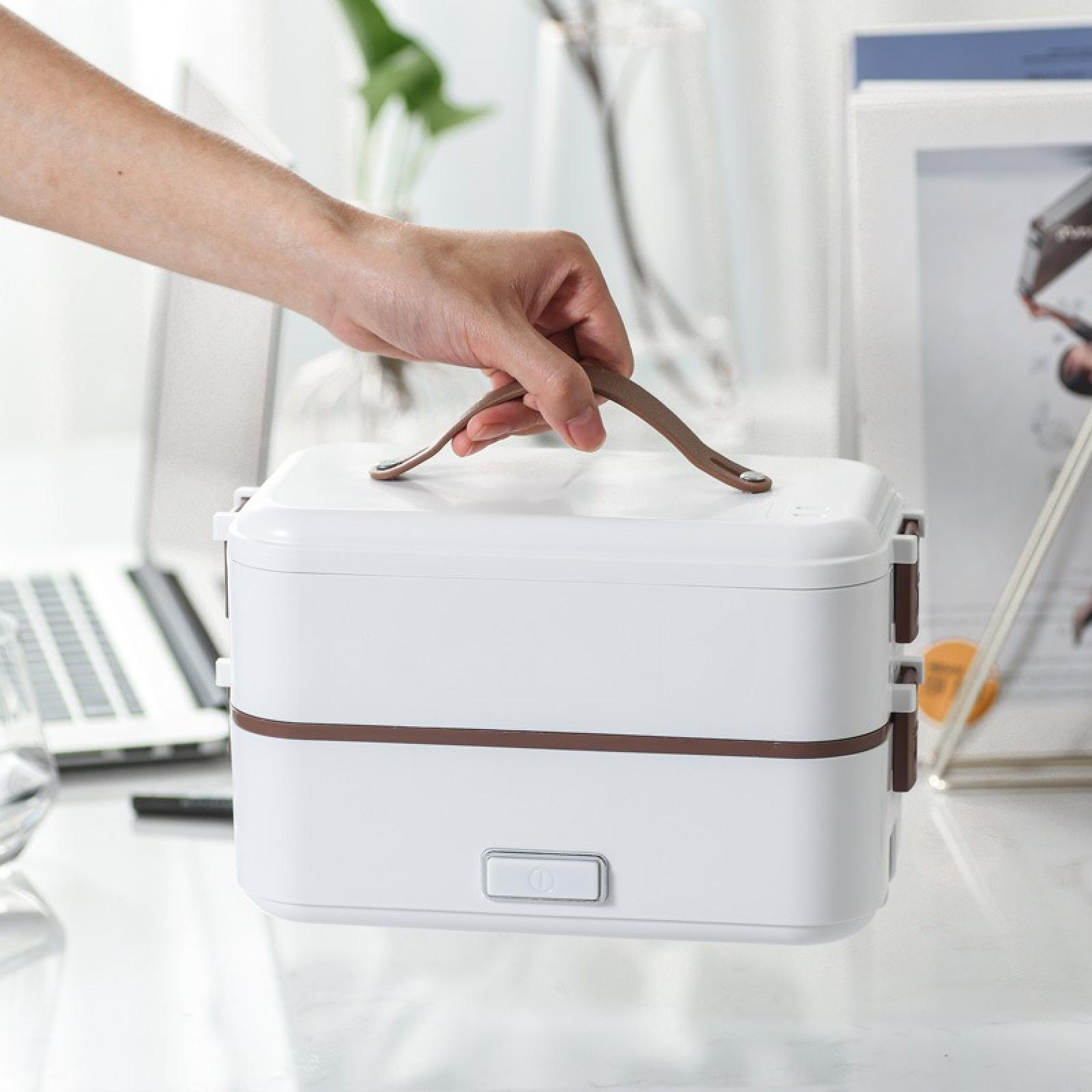Hand hält eine weiße Lunchbox mit braunen Akzenten und Griff. Ein Laptop, eine Pflanze und andere Gegenstände sind im Hintergrund.