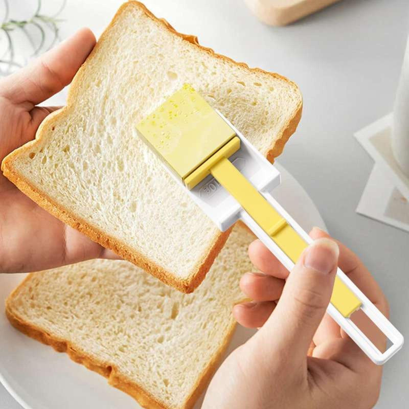 Eine Hand hält eine Brotscheibe und trägt Butter mit einem gelb-weißen Streuer auf. Eine weitere Scheibe liegt auf einem Teller.