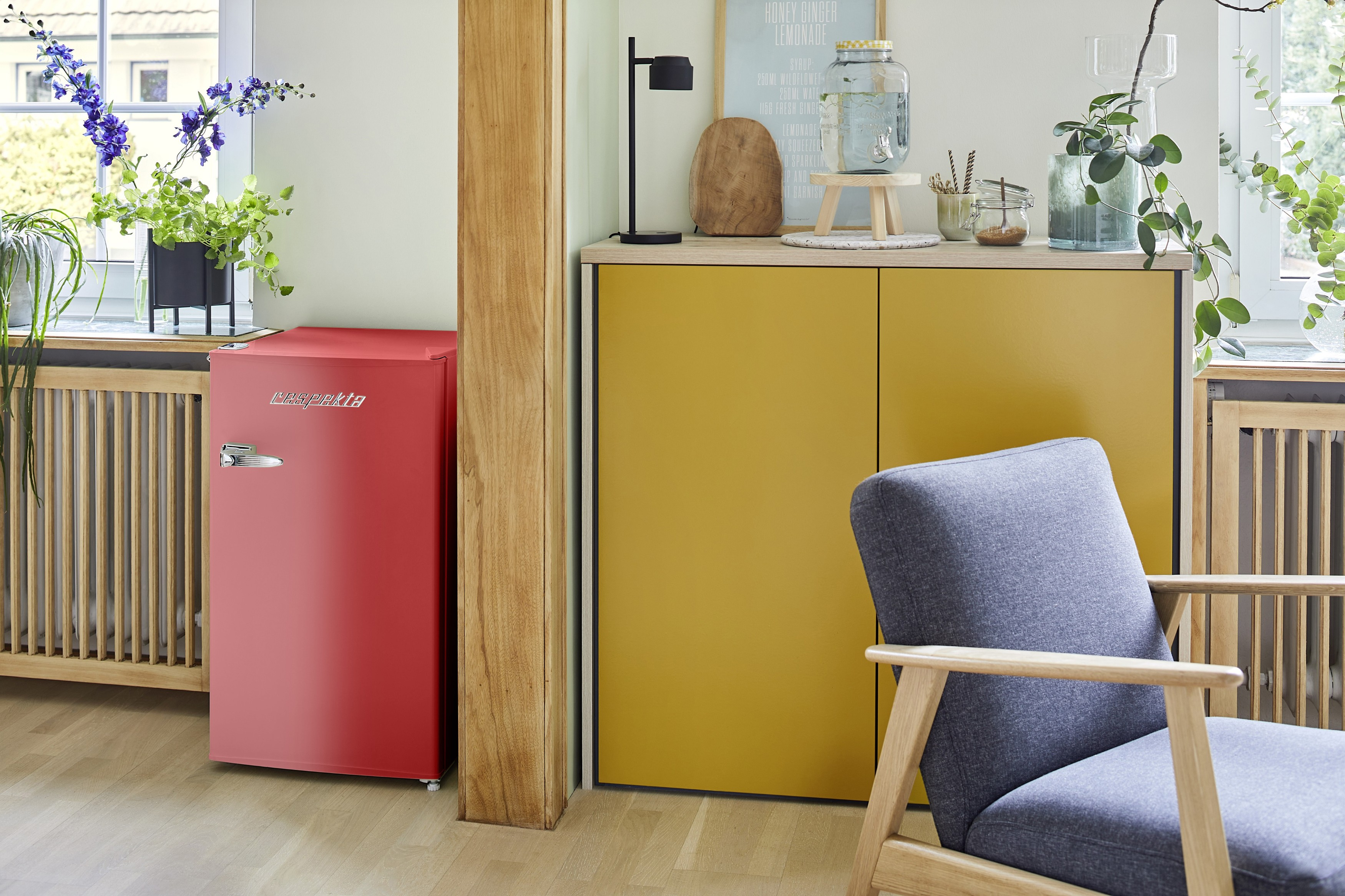 Ein roter Kühlschrank, ein gelber Schrank, ein grauer Stuhl und Pflanzen in einem Raum mit Fenster.