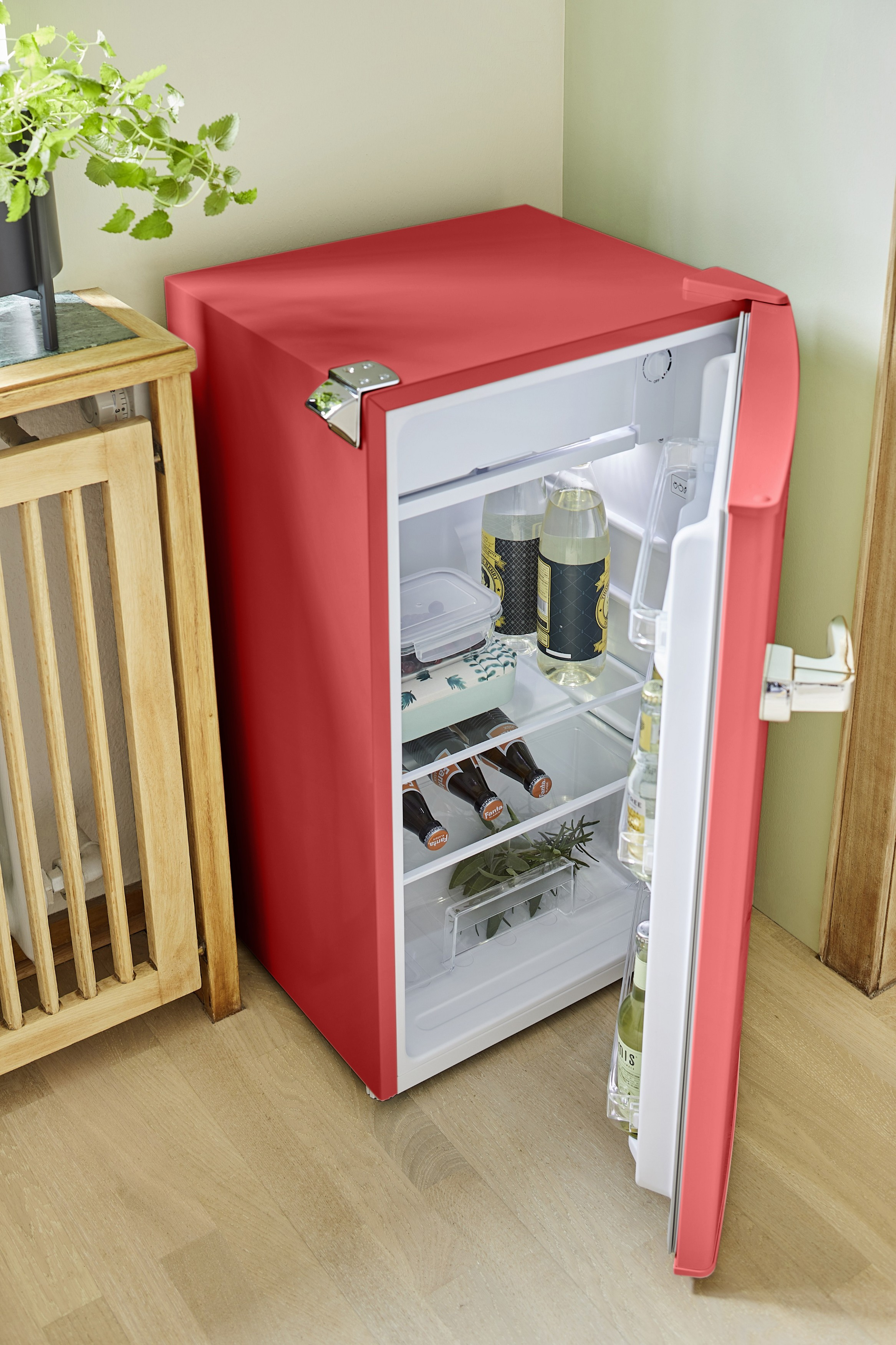 Ein roter Mini-Kühlschrank ist geöffnet und zeigt Getränke im Inneren. Ein Holztisch und eine Pflanze sind in der Nähe.