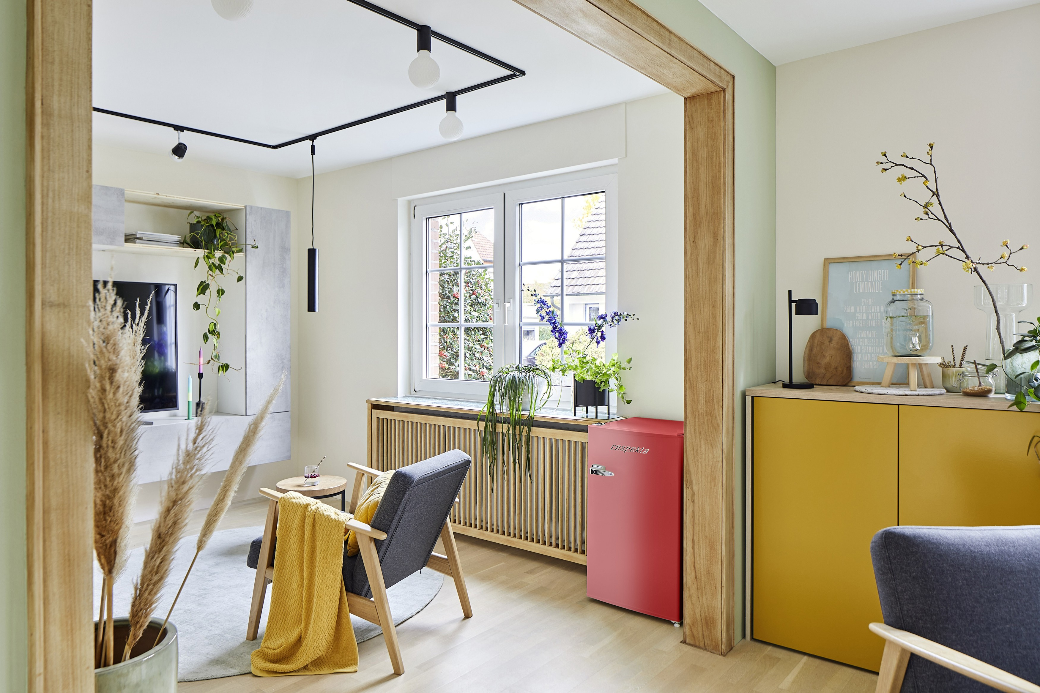 Wohnzimmer: grauer Stuhl mit gelber Decke, roter Kühlschrank, gelber Schrank, Fenster mit Pflanzen, TV und Dekoration.