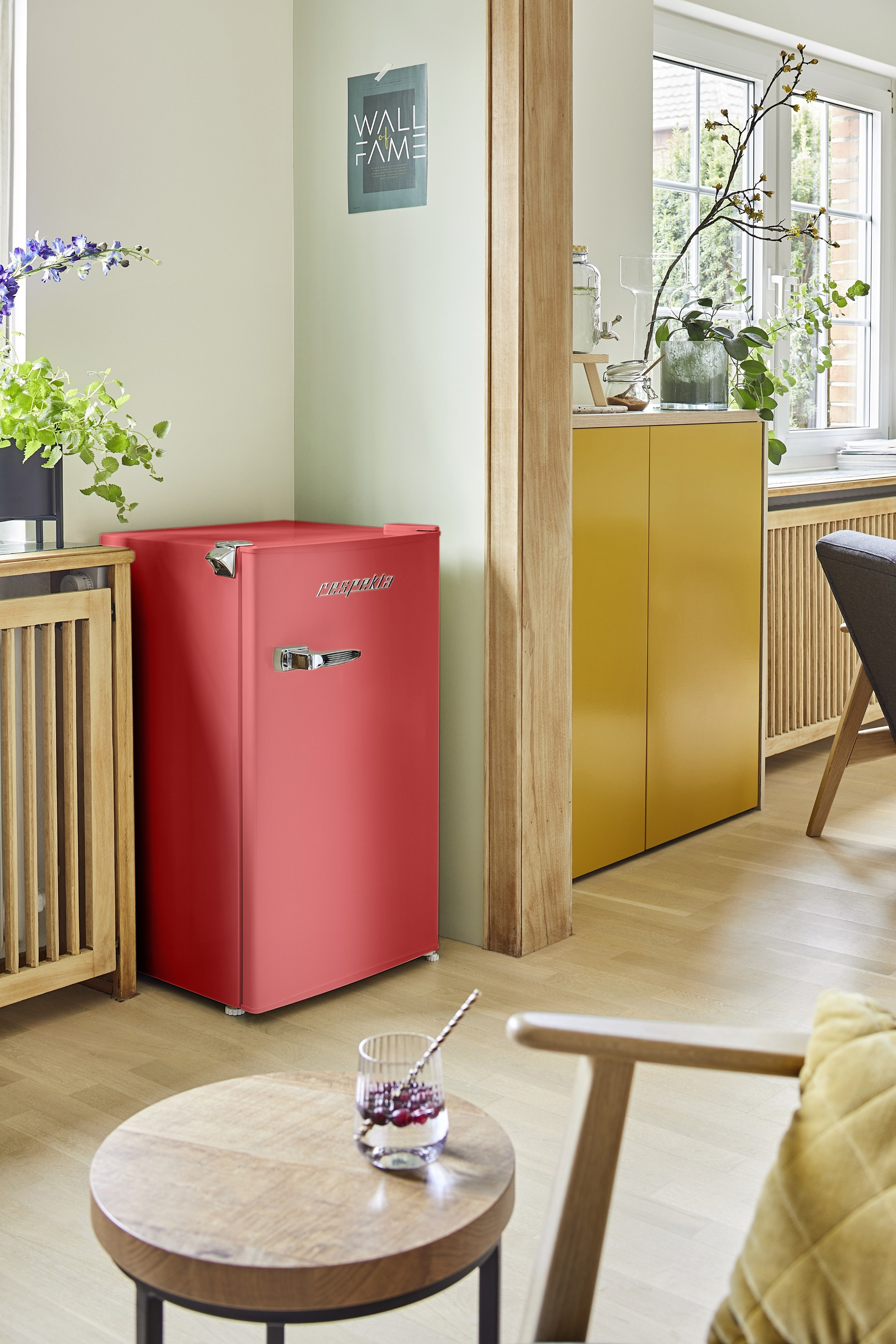 Ein roter Kühlschrank steht neben einem gelben Schrank. Ein Glas Getränk steht auf einem Holztisch. Im Hintergrund ein Fenster.