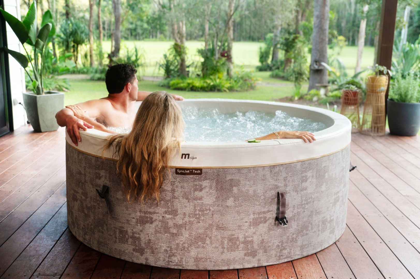 Ein Paar entspannt sich in einem Whirlpool. Die Frau hat lange blonde Haare. Der Whirlpool steht auf einem Holzdeck mit Grün im Hintergrund.