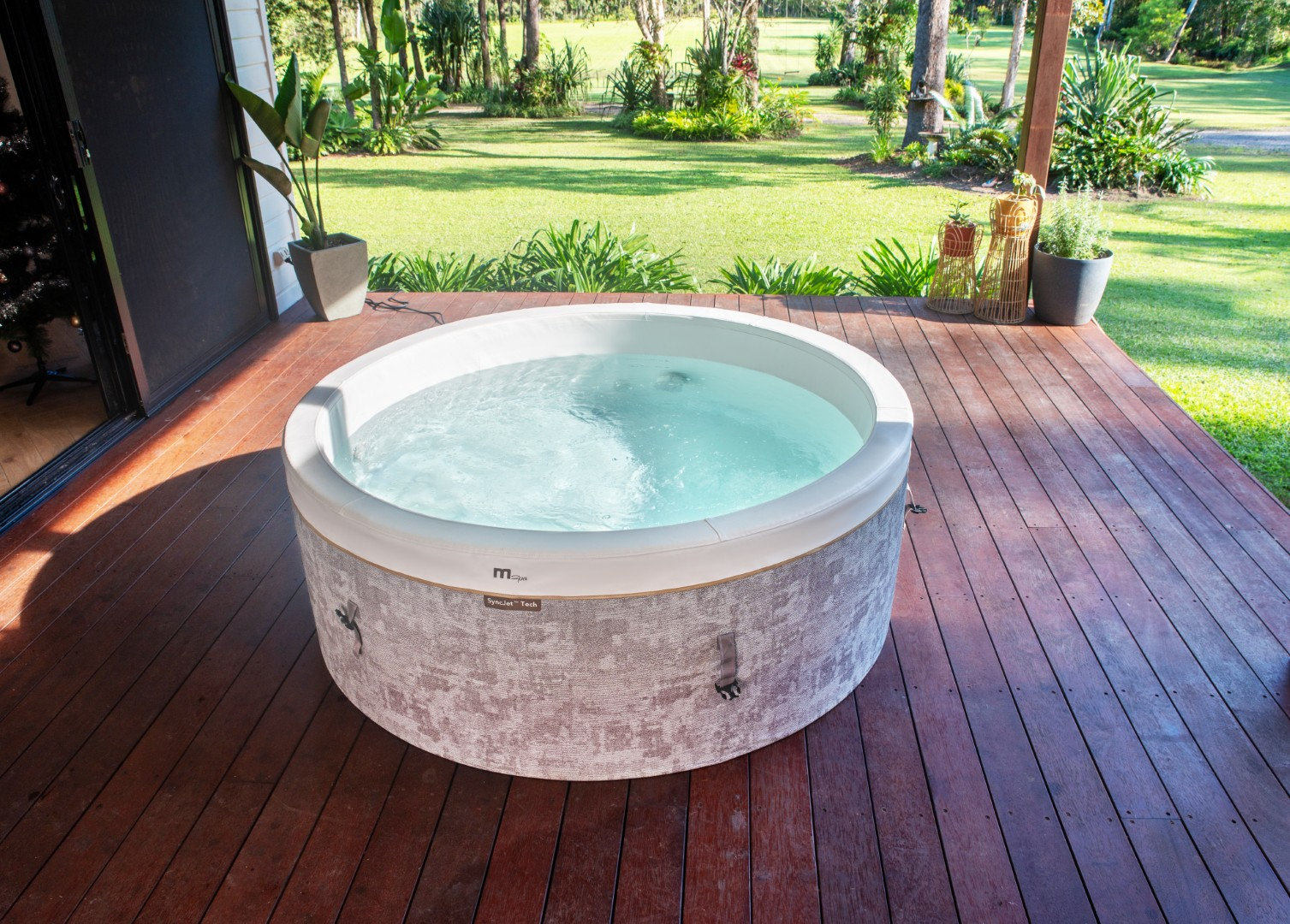Ein runder Whirlpool mit klarem Wasser steht auf einer Holzterrasse. Im Hintergrund sind grünes Gras und Bäume zu sehen.