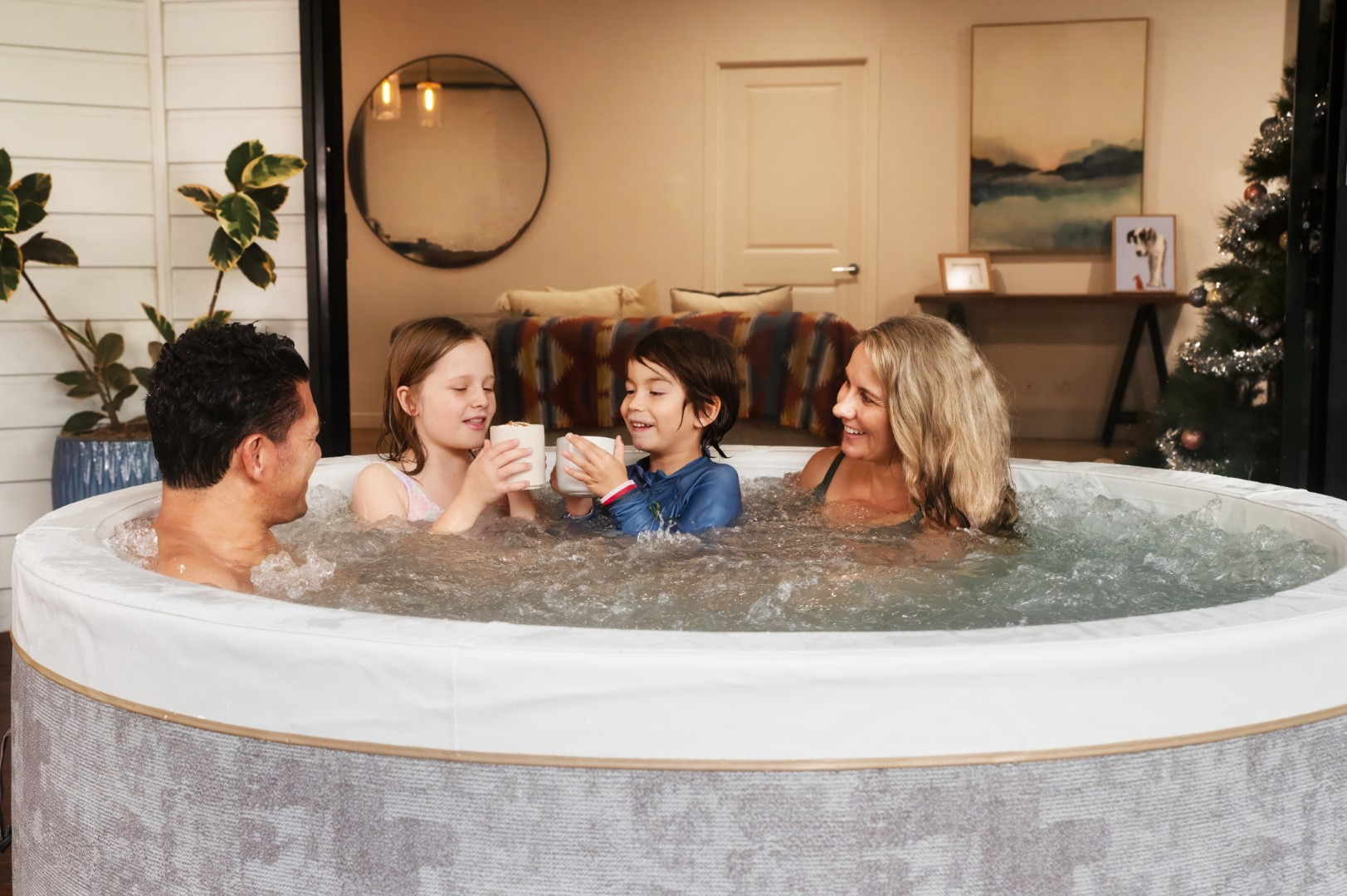 Eine vierköpfige Familie in einem Whirlpool, die Tassen hält. Sie lächeln. Der Hintergrund zeigt ein Wohnzimmer und einen Weihnachtsbaum.