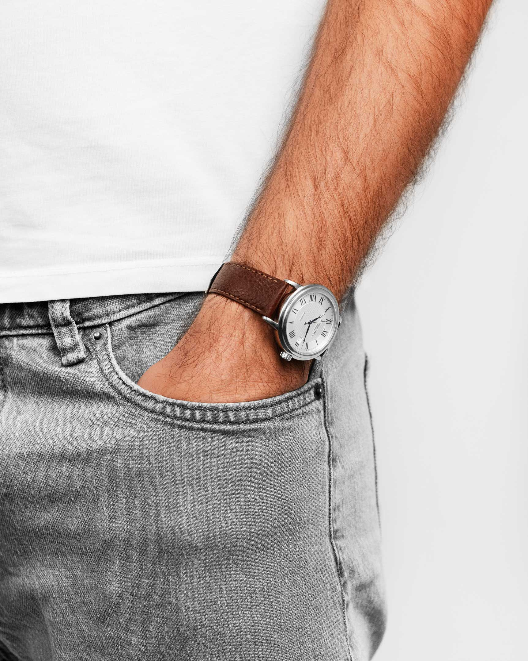 Ein Arm mit Uhr. Er trägt eine braune Lederuhr und graue Jeans. Seine Hand steckt in der Tasche.