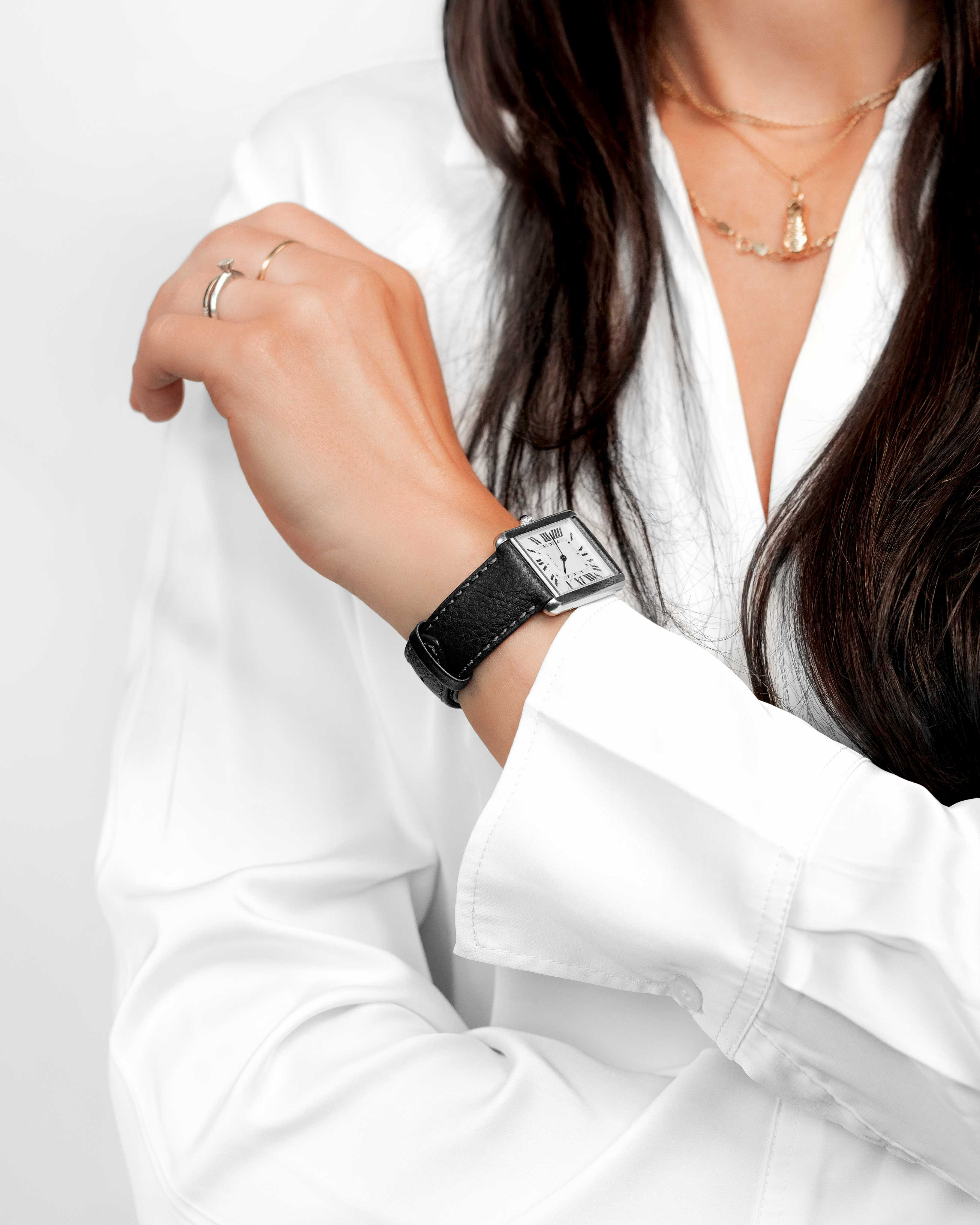 Eine Person trägt ein weißes Hemd und eine schwarze Uhr. Die Hand der Person ist sichtbar. Eine goldene Halskette ist ebenfalls sichtbar.