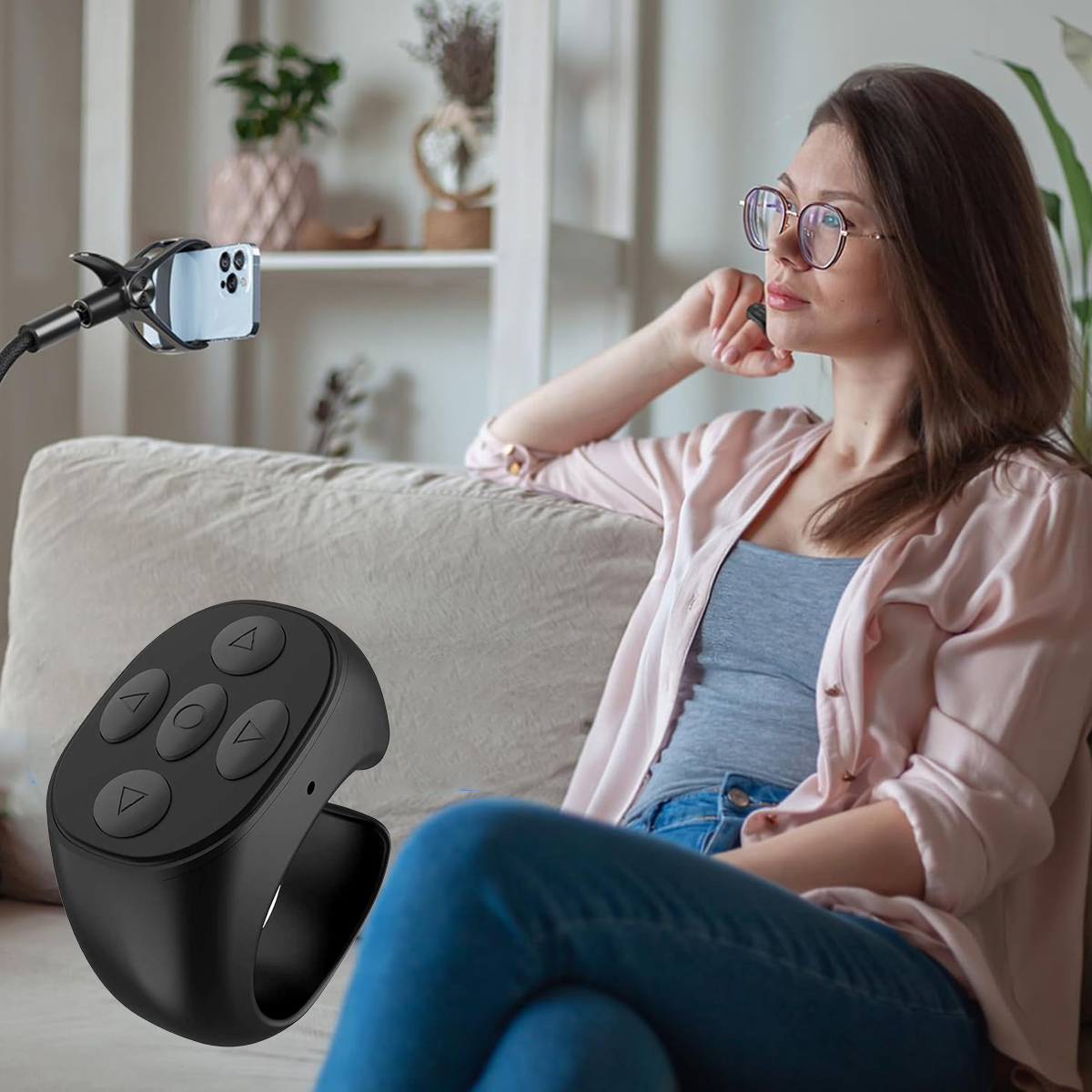 Frau mit Brille sitzt neben Handy auf einem Ständer. Schwarze Ringfernbedienung im Vordergrund. Hintergrund: Sofa und Regale.