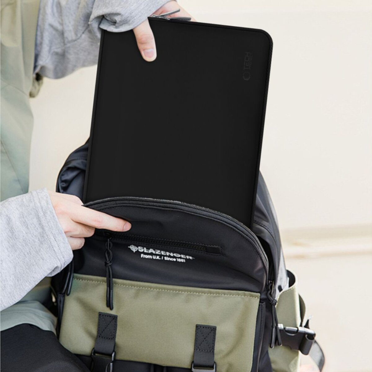 Een persoon plaatst een zwarte laptoptas in een rugzak. De rugzak is zwart en olijfgroen.
