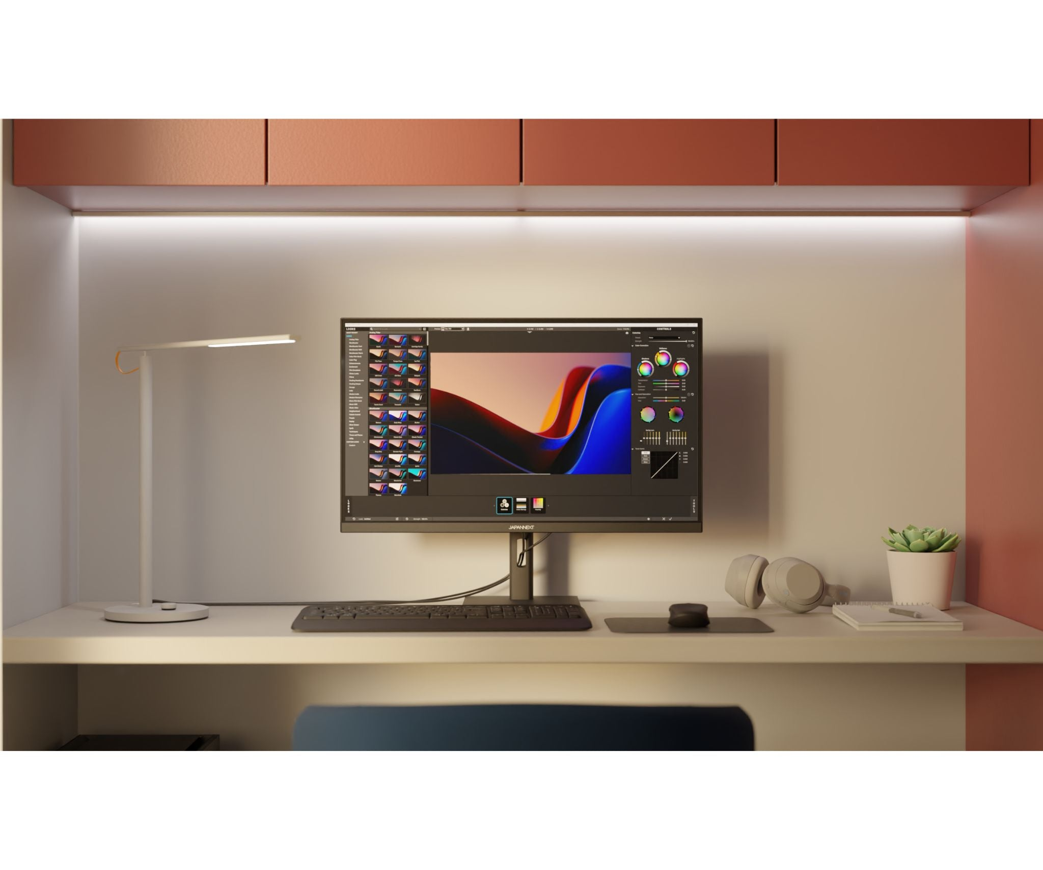 Una configuración de escritorio con un monitor que muestra software de edición, una lámpara, auriculares y una planta. El fondo es melocotón.